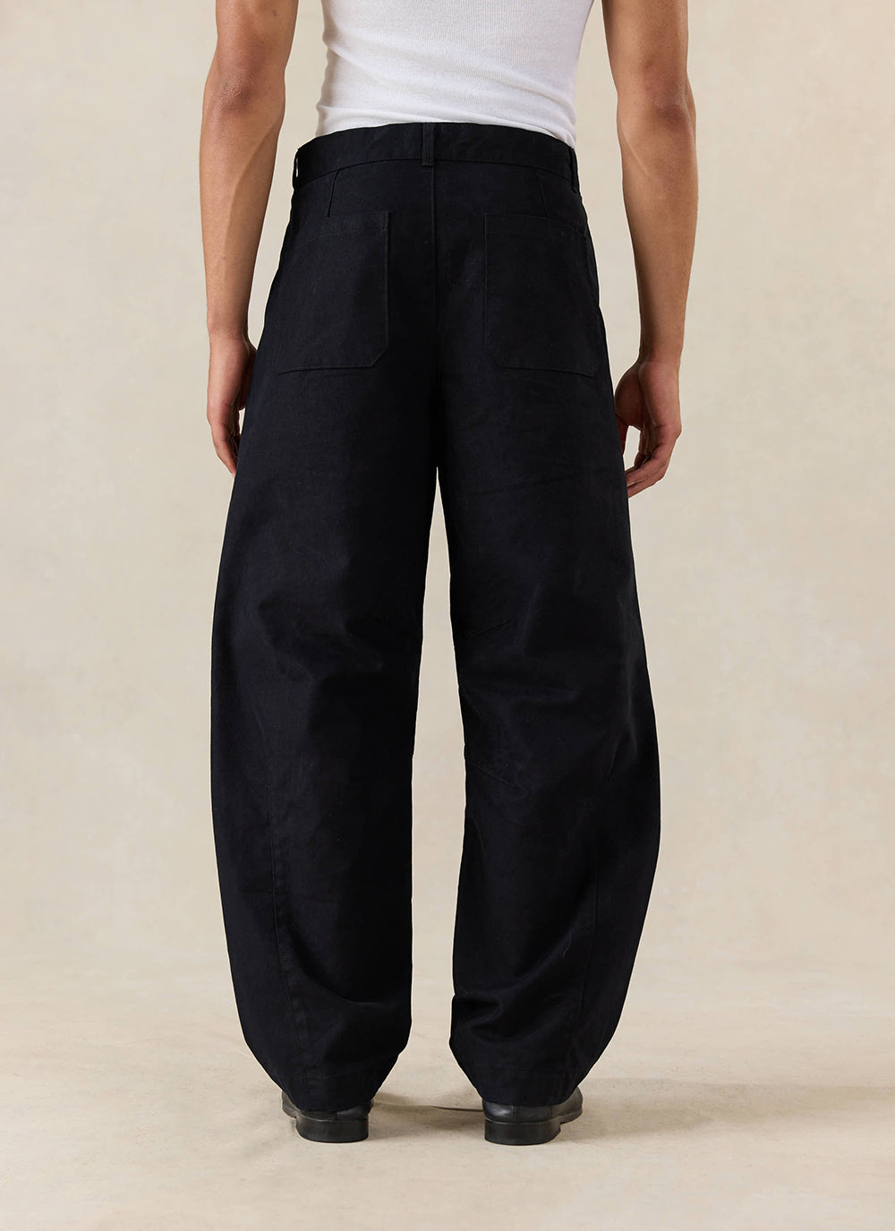 A model wears the Percival Milo Relaxed Barrel Leg Trousers in black, shown from the rear and styled with a white vest.