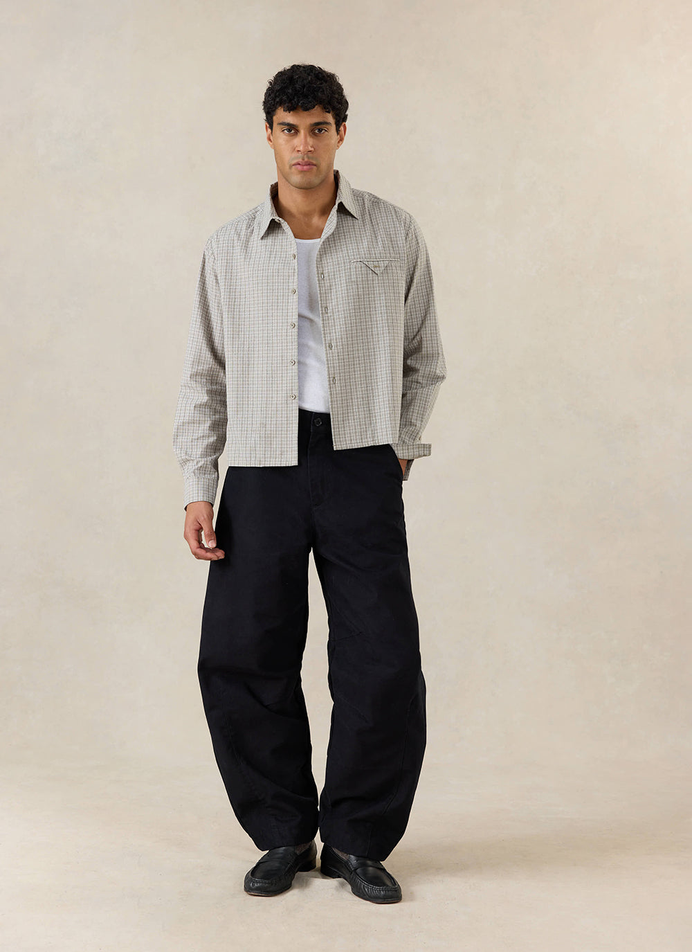 A model wears the black Percival Milo Relaxed Barrel Leg Trousers, styled with a checkered shirt and white tank top.