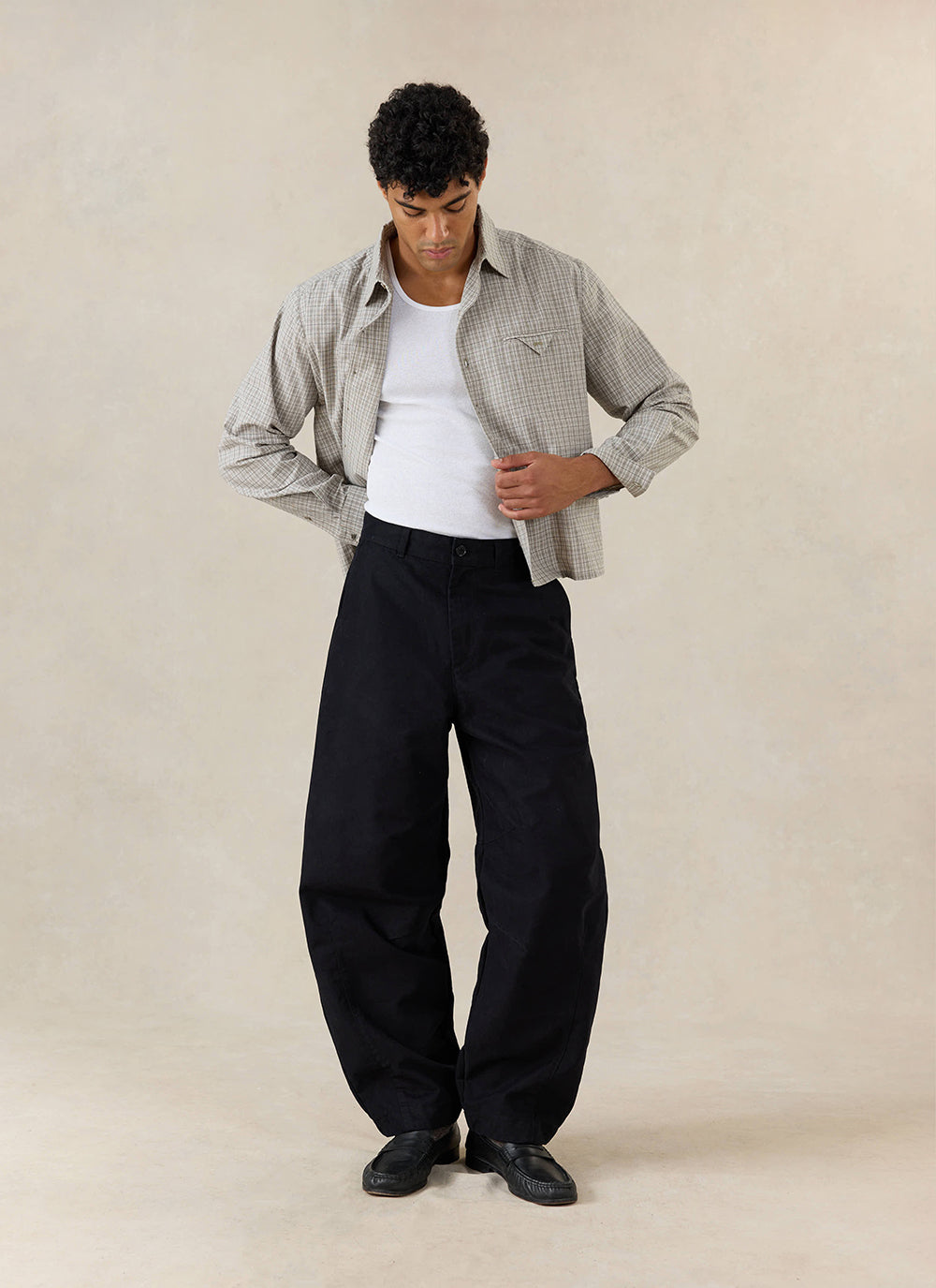A model wears black Percival Milo Relaxed Barrel Leg Trousers, a check shirt and white vest in a studio.
