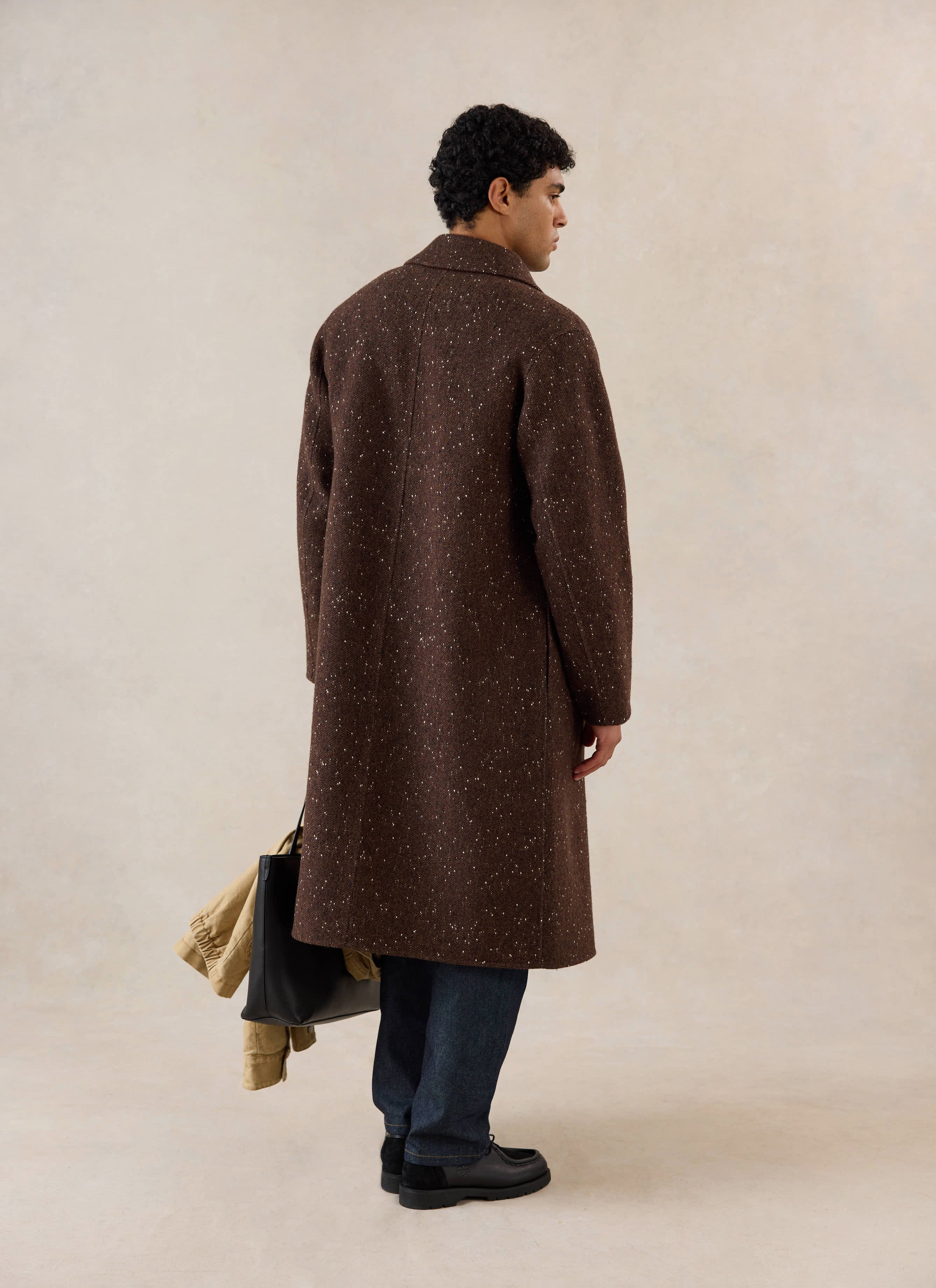 A rear view of a model wearing the brown fleck Percival Minnesota overcoat, carrying a black tote bag.