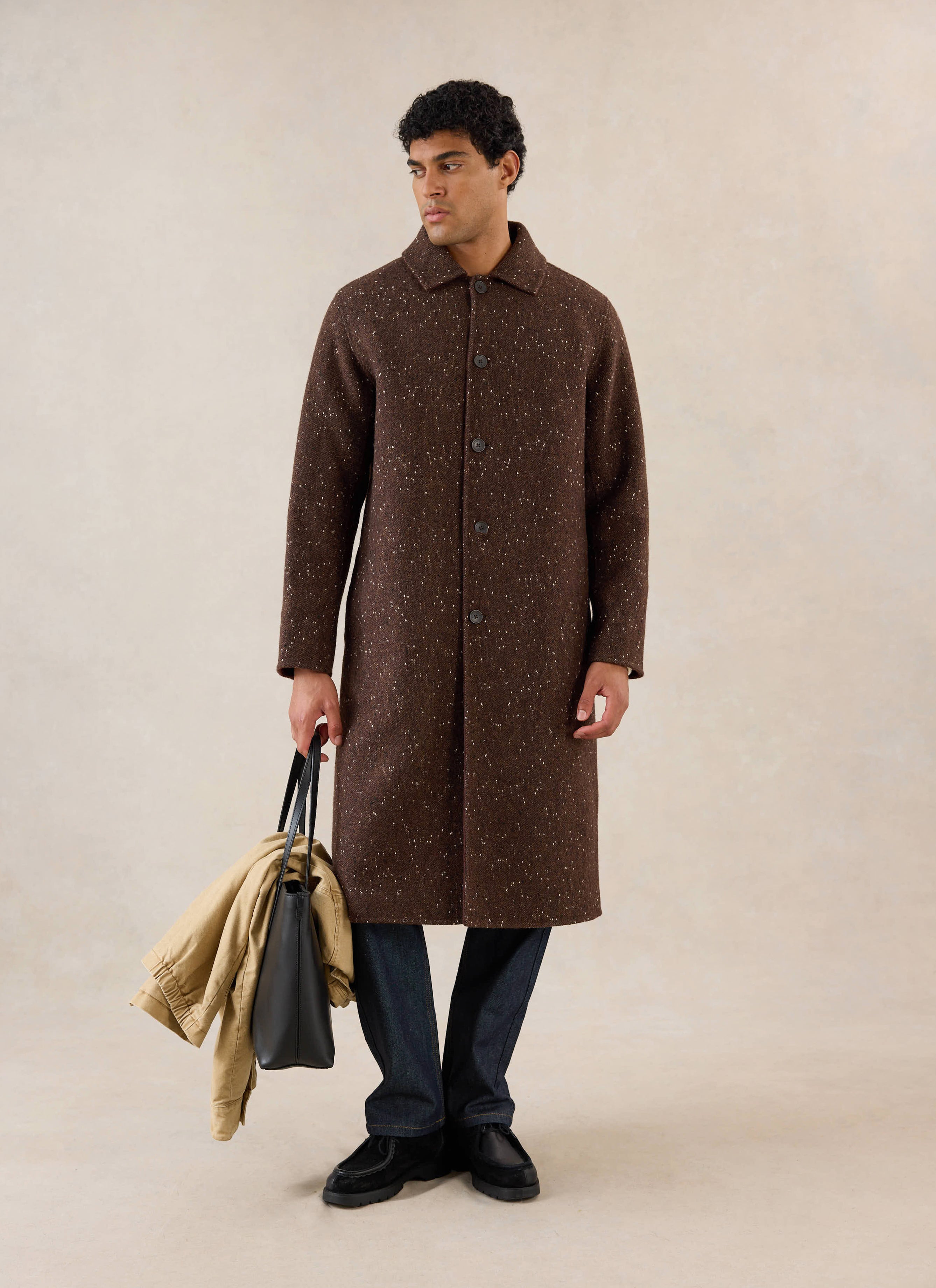A model stands wearing the brown fleck Minnesota Overcoat from Percival, holding a black leather tote bag in a studio.