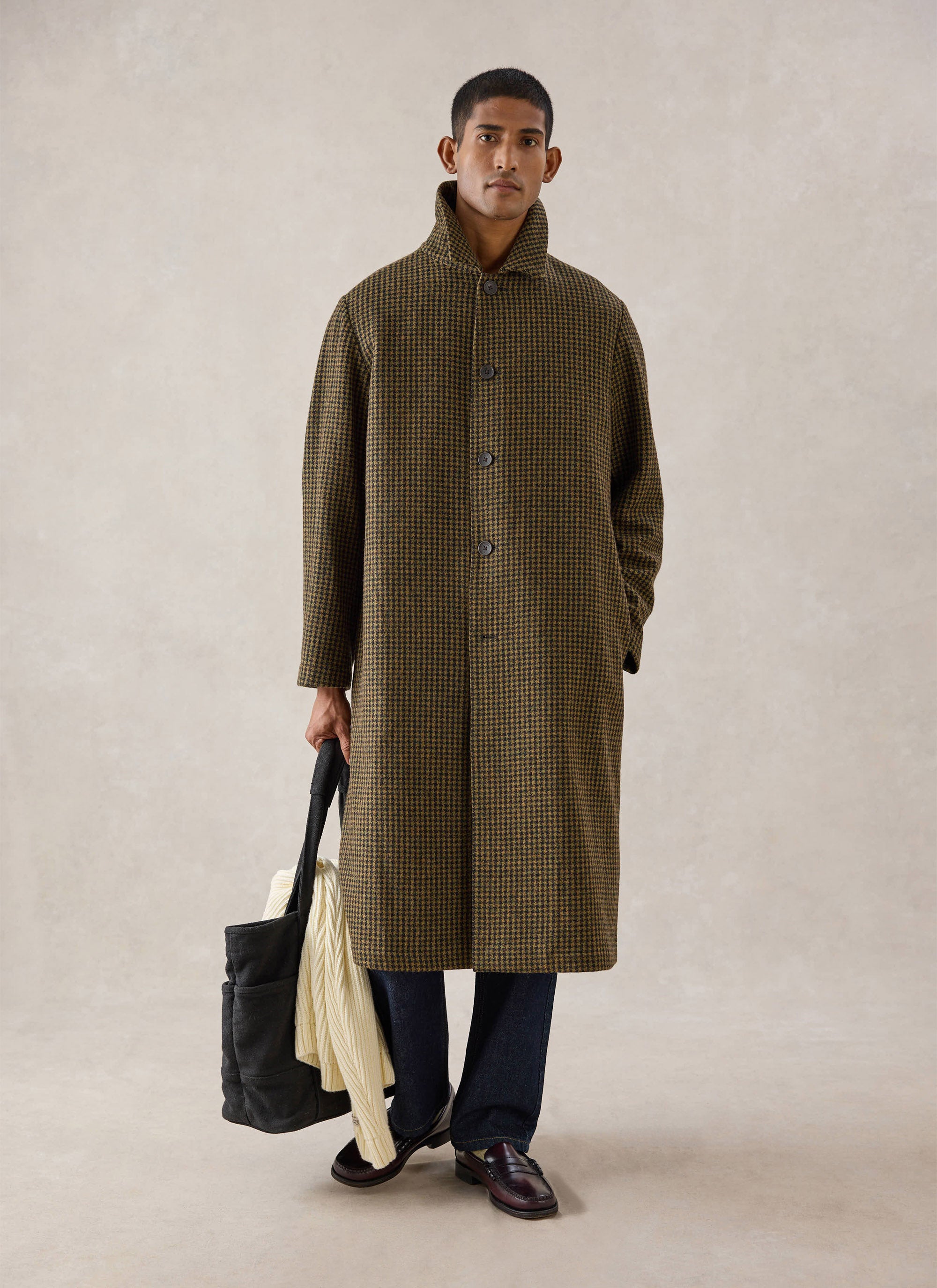 A model stands in a studio wearing the Watcher Check Minnesota Overcoat from Percival, holding a dark bag and knit jumper.