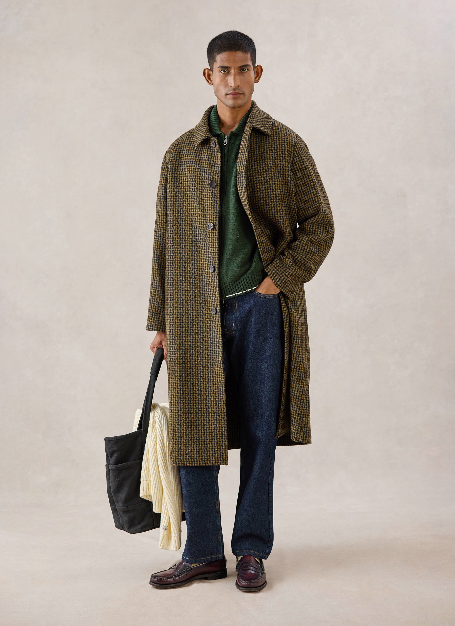 A model wears the Percival Minnesota Overcoat in watcher check over a green zip knit and dark trousers in a studio.