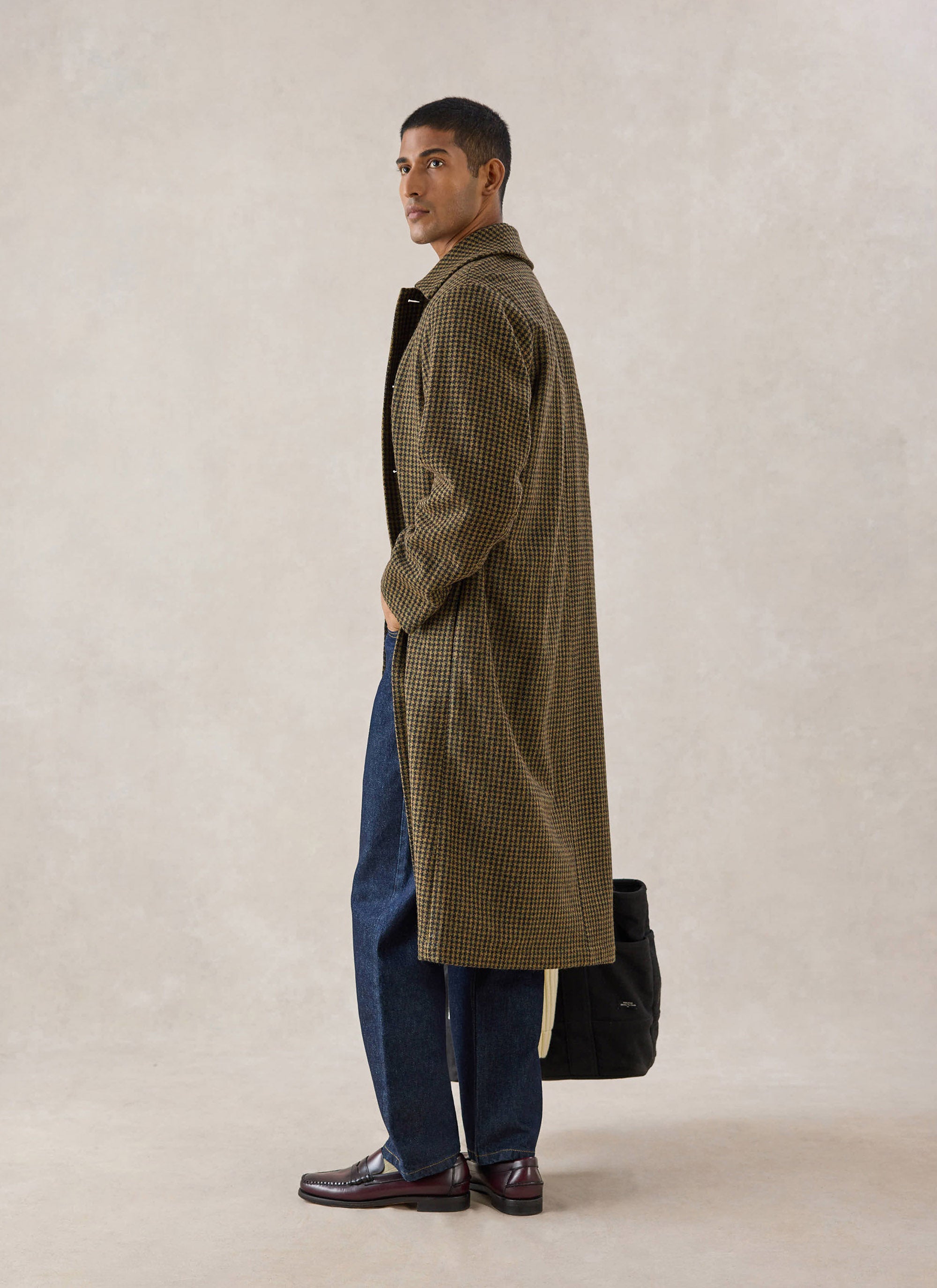A model wears the watcher check Percival Minnesota overcoat with blue jeans and loafers, holding a black bag.