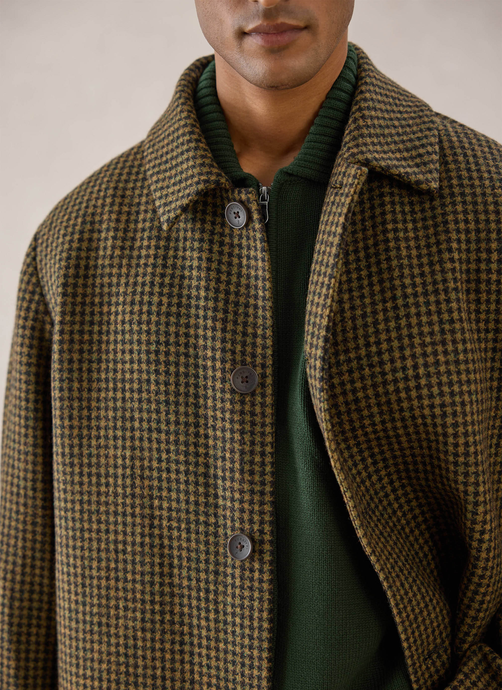 A close-up of a model wearing the watcher check Minnesota overcoat from Percival over a green knit jumper.
