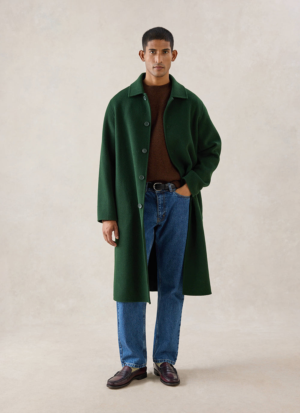 A model wears the forest green wool Percival Minnesota Overcoat with a brown jumper and blue jeans in a studio.