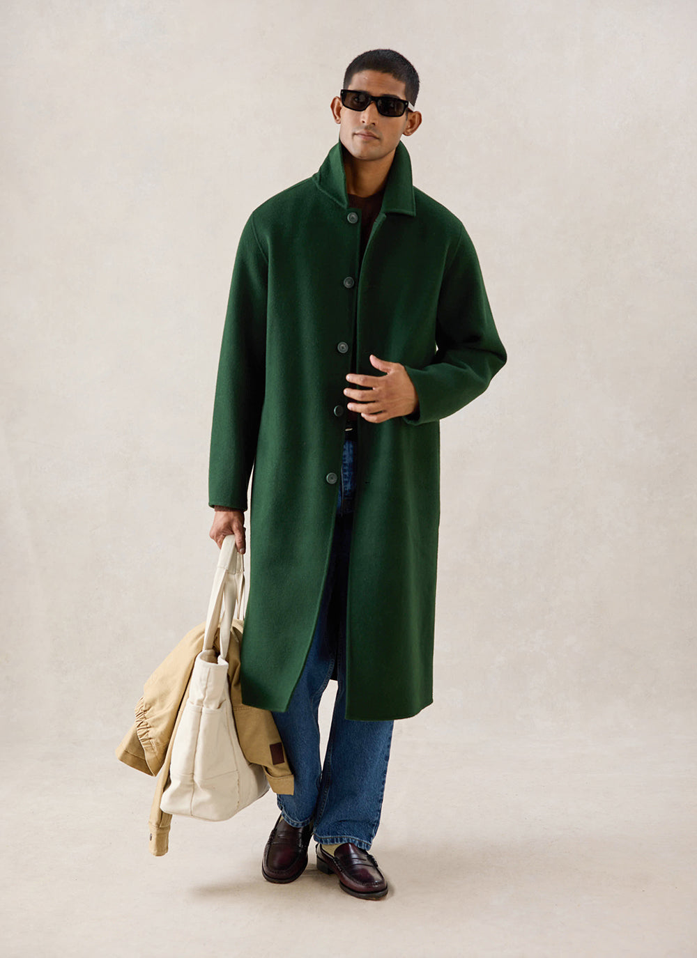 A model wears the forest green wool Percival Minnesota Overcoat, styled with blue jeans and holding a cream tote bag.