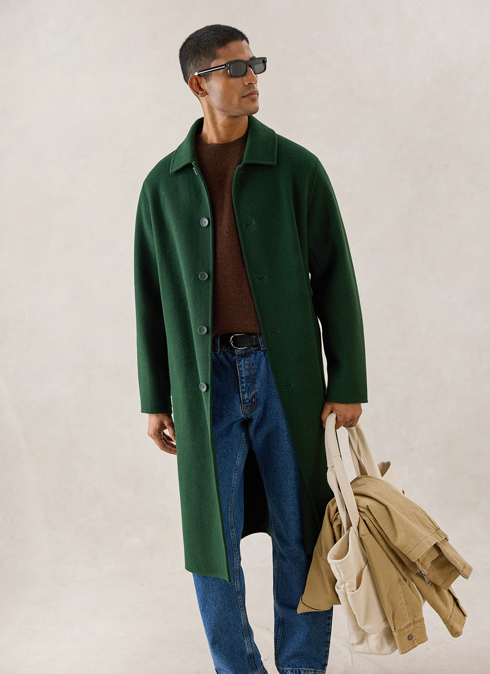 A model wears the forest wool Percival Minnesota Overcoat, styled with a brown jumper and jeans, holding a tote bag.