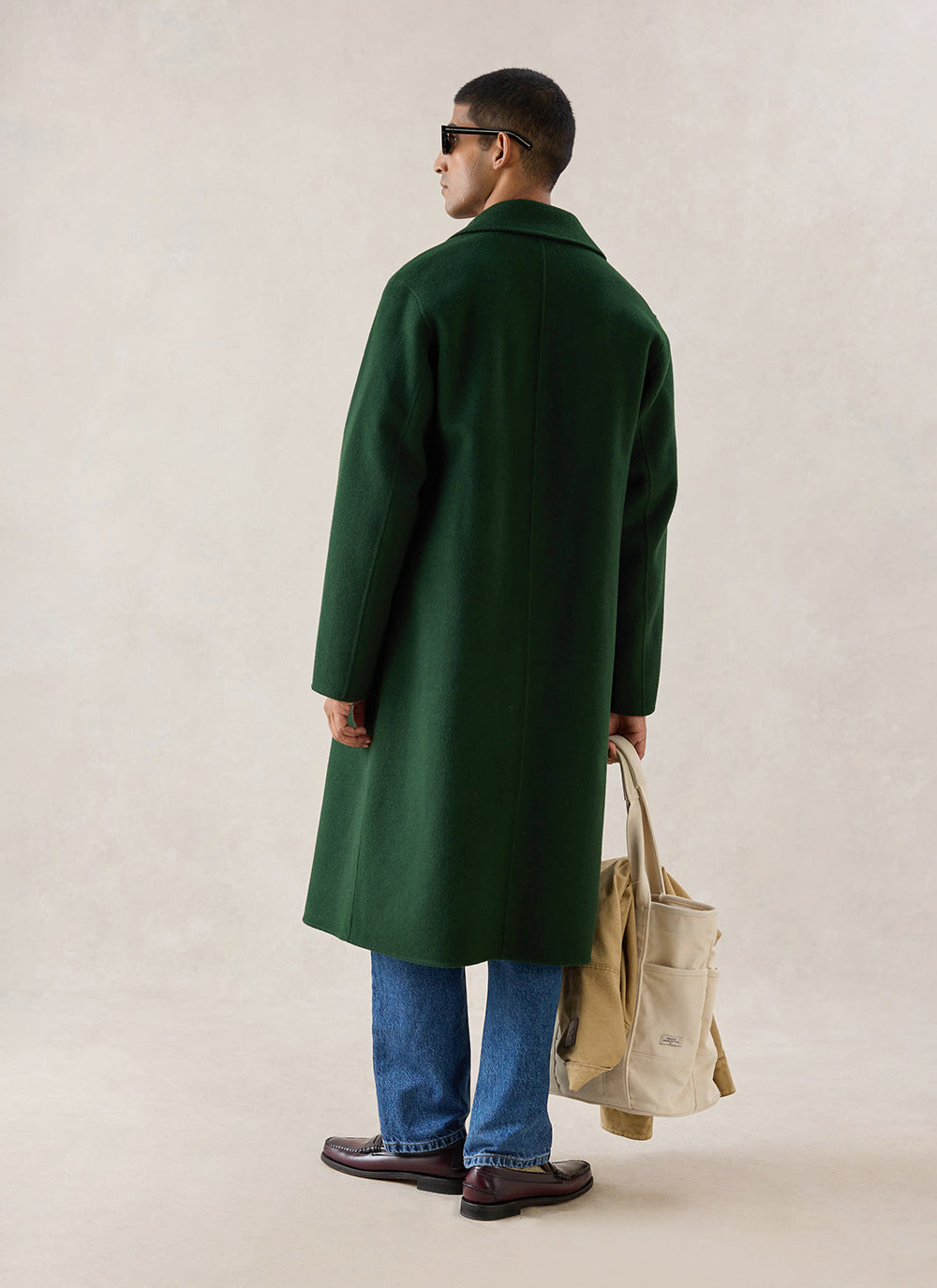 A model from behind wears the forest green wool Percival Minnesota Overcoat, styled with blue jeans and carrying a tote bag.