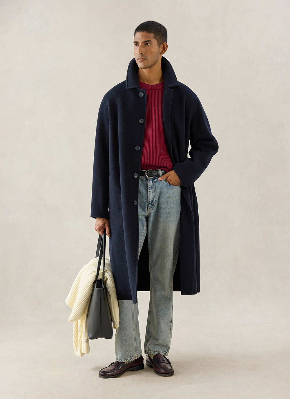 A model wears the navy wool Minnesota Overcoat from Percival, styled with a red knit jumper, jeans and a tote bag.