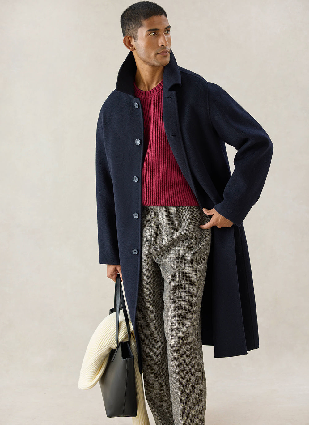 A model wears the navy wool Percival Minnesota Overcoat with a red jumper and patterned trousers, holding a bag.