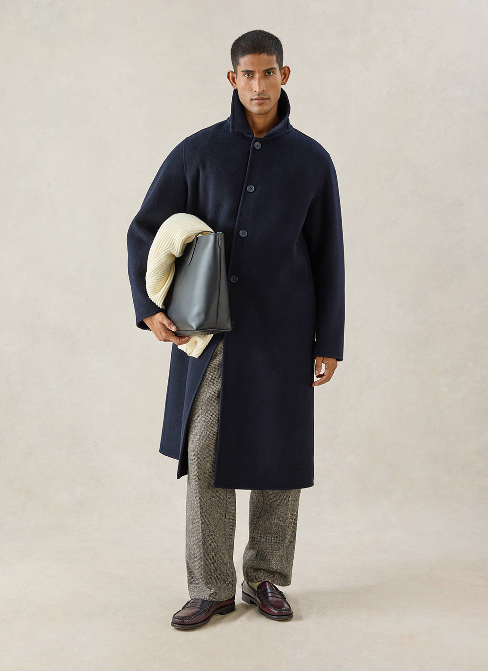 A model wears the navy wool Percival Minnesota Overcoat, holding a dark bag and cream knit, with patterned trousers.