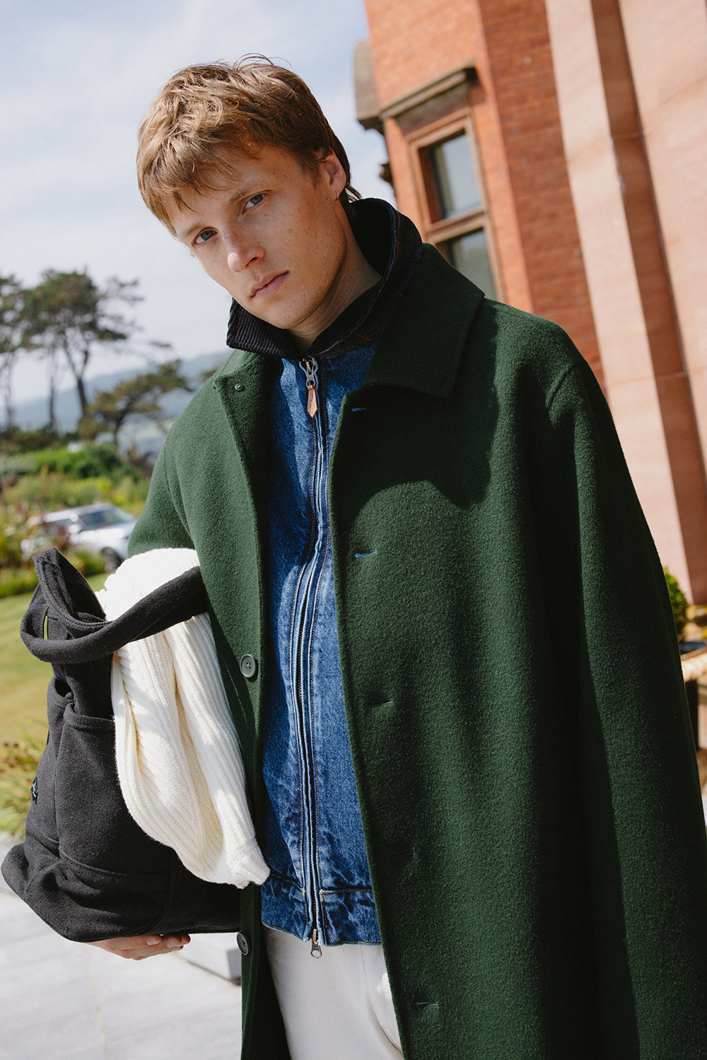 A model wears the Percival Minnesota double-faced wool overcoat in forest green over a blue zip jacket, holding a dark bag.