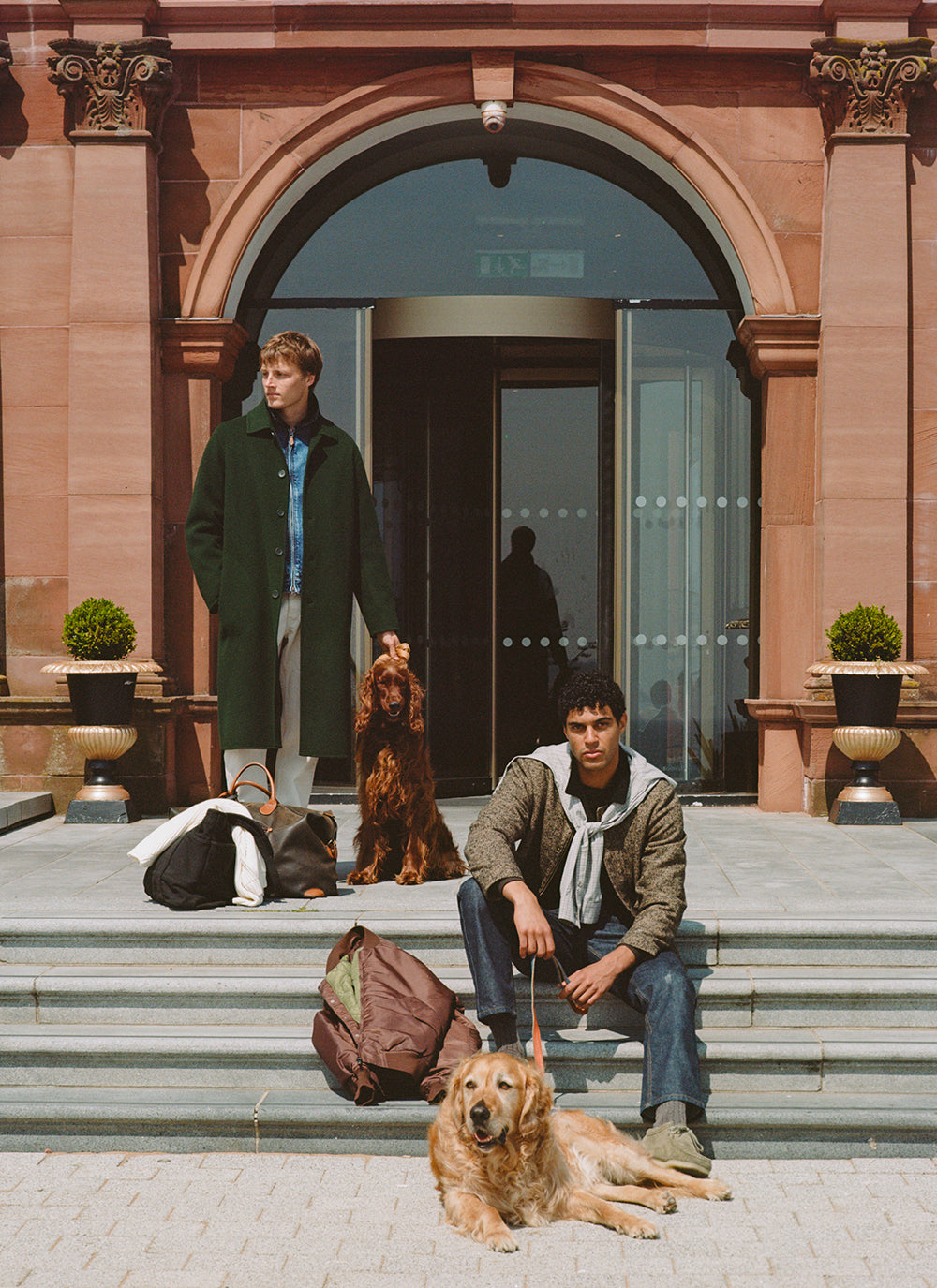 Two models and two dogs outside a building, with one wearing a green Percival wool overcoat and the other a patterned jacket.