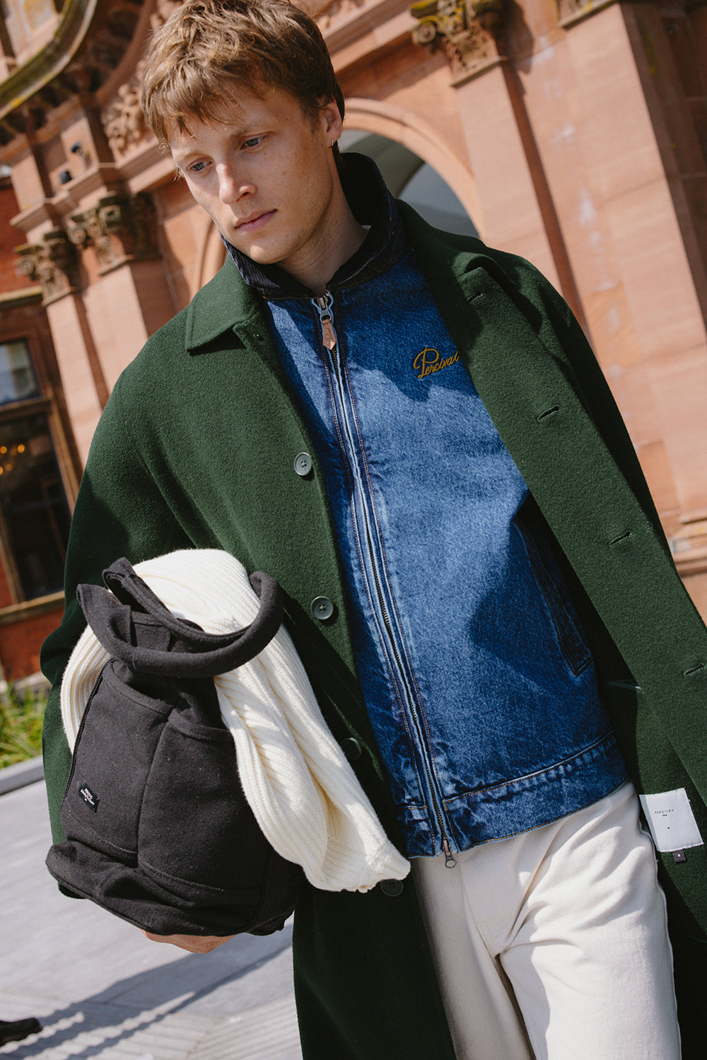 A model wears a Percival mid wash blue denim zip jacket under a green coat, carrying a dark bag.