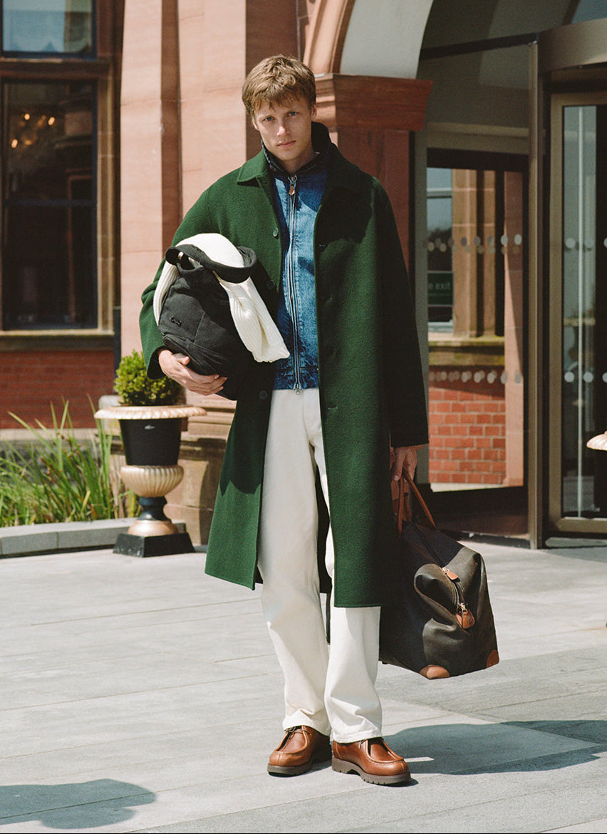 A model wearing a forest green Percival wool overcoat over a denim jacket and cream trousers outside an entrance.