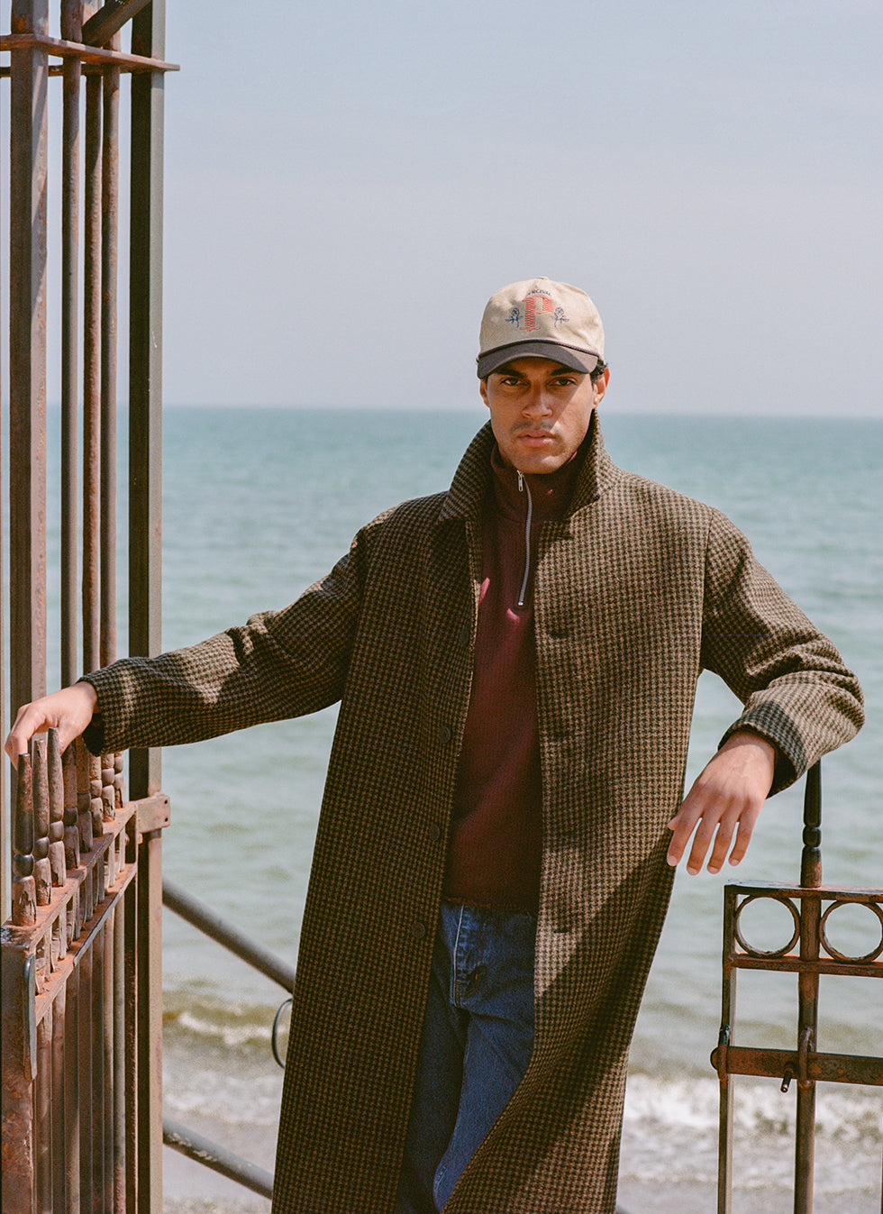 A model wearing the Percival Minnesota wool overcoat in Watcher Check over a burgundy zip jumper, cap, and jeans by the sea.