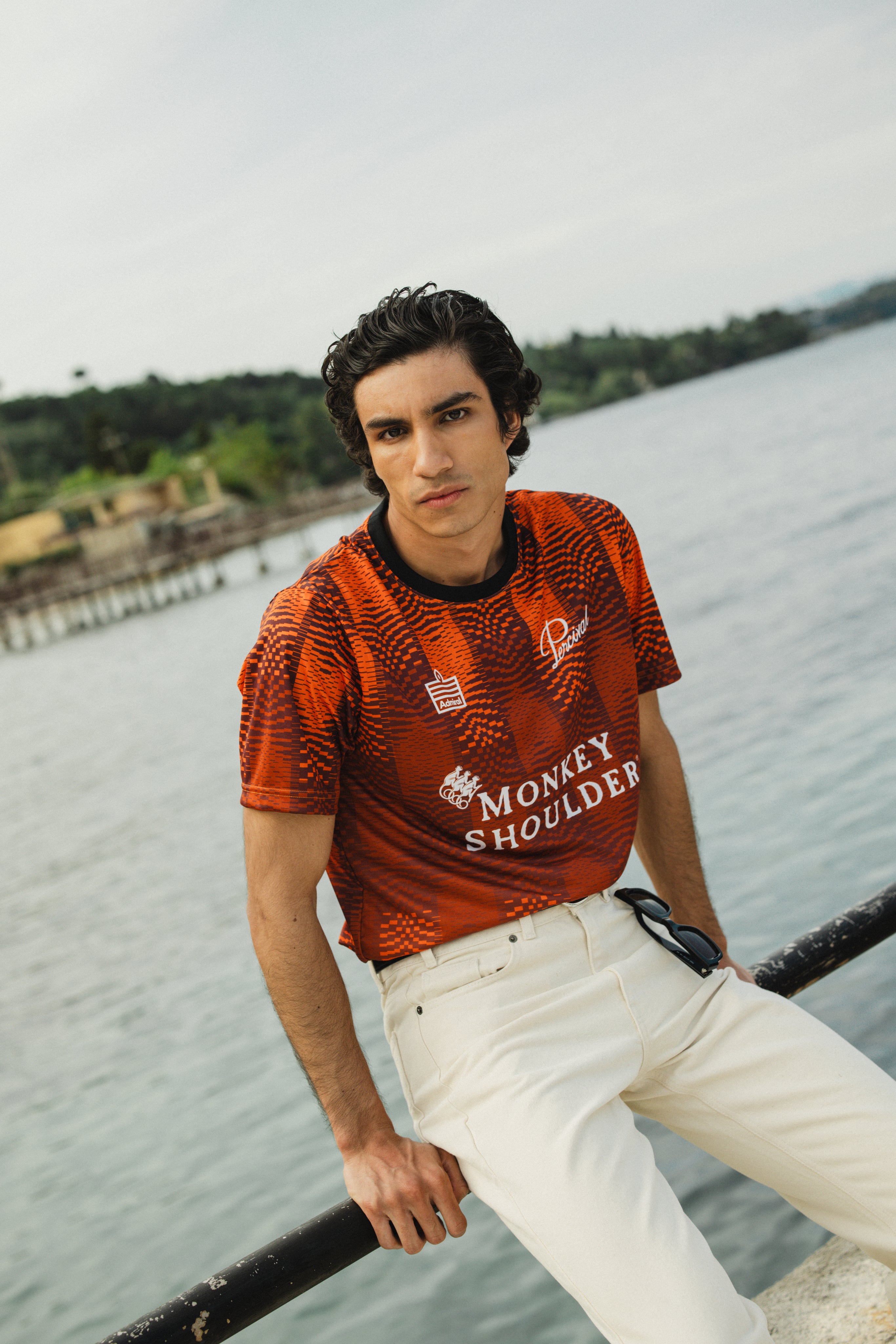 Orange Percival x Monkey Shoulder football shirt featuring pixel-distortion stripes, worn by a model by the water.