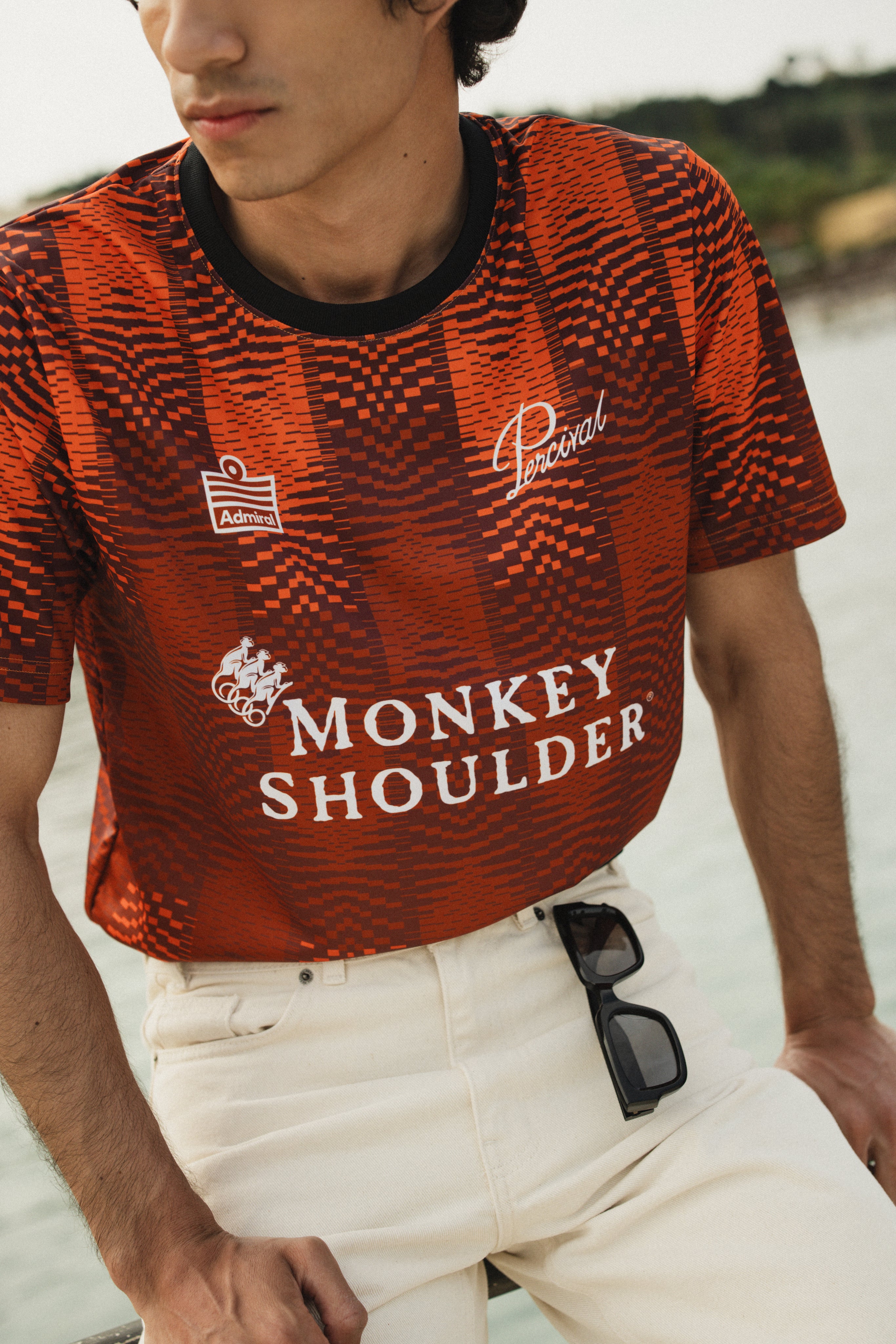 The Percival x Monkey Shoulder orange football shirt with pixel-distortion stripes, worn by a model in white trousers.