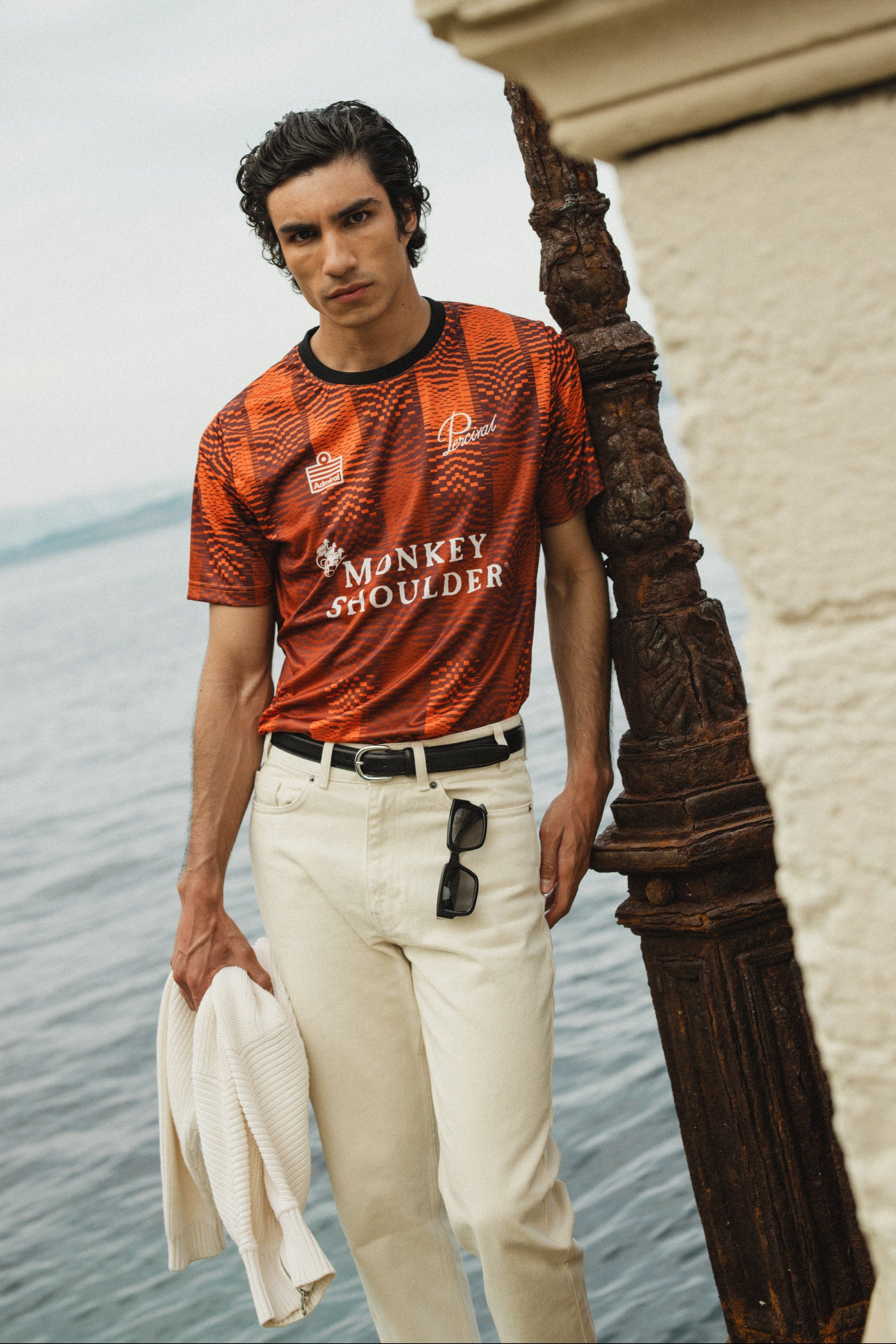 The Admiral x Percival x Monkey Shoulder orange pixel-distortion football shirt, styled with white trousers by the sea.