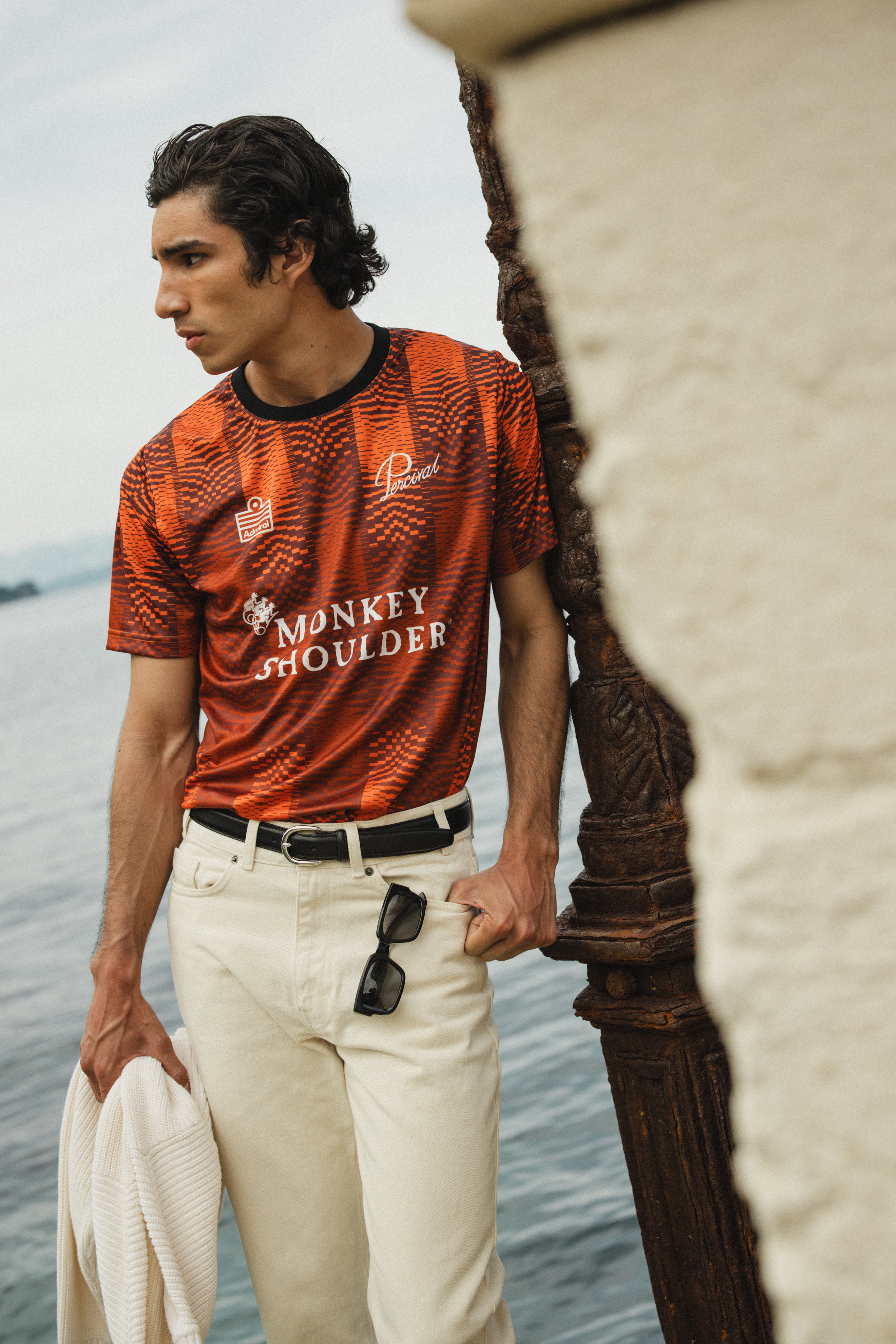 The orange Percival x Monkey Shoulder football shirt with a pixel-distortion stripe pattern, worn by a model near the coast.
