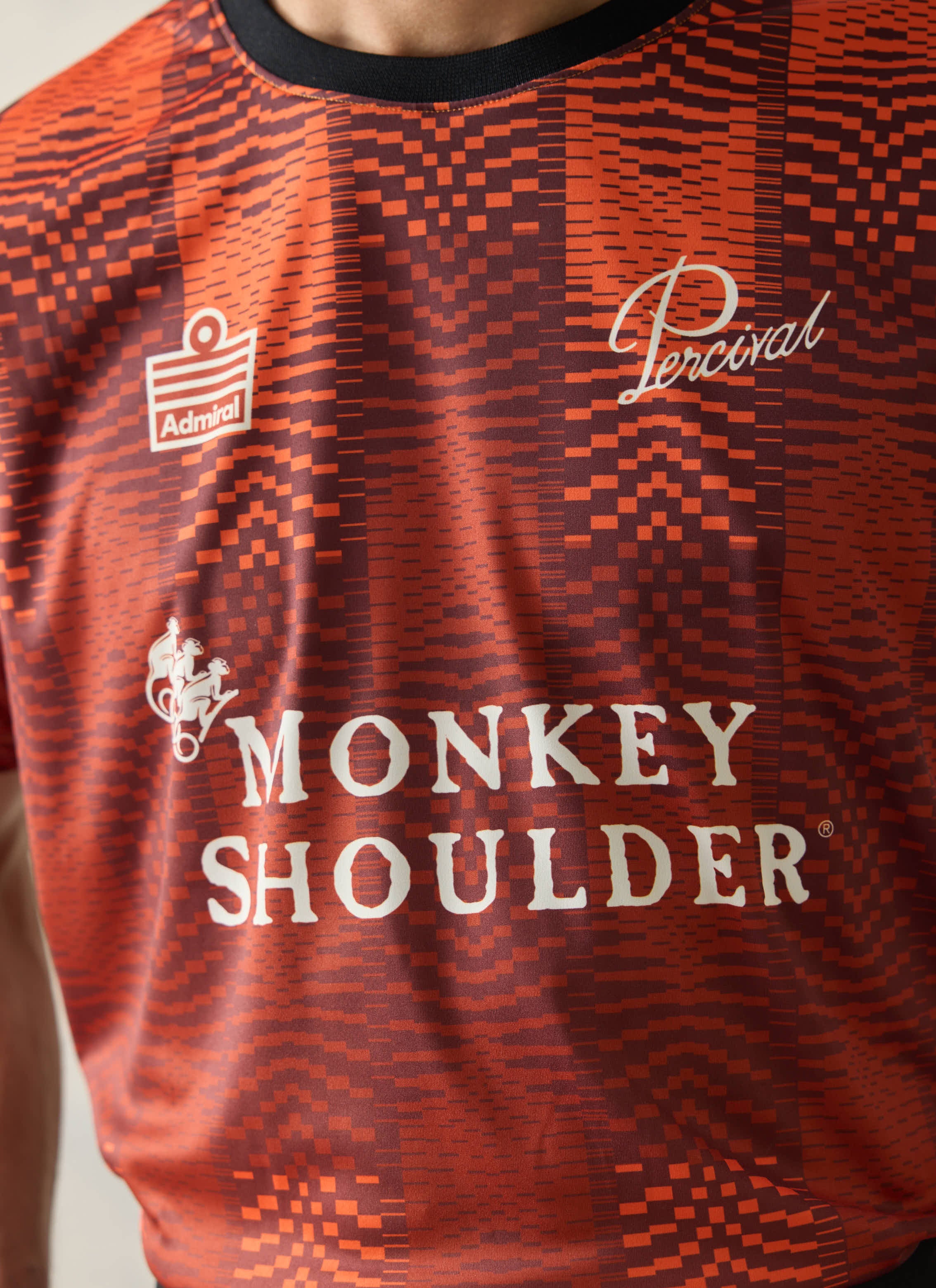 Orange Percival x Monkey Shoulder football shirt featuring a pixel-distortion stripe pattern and white logo branding.
