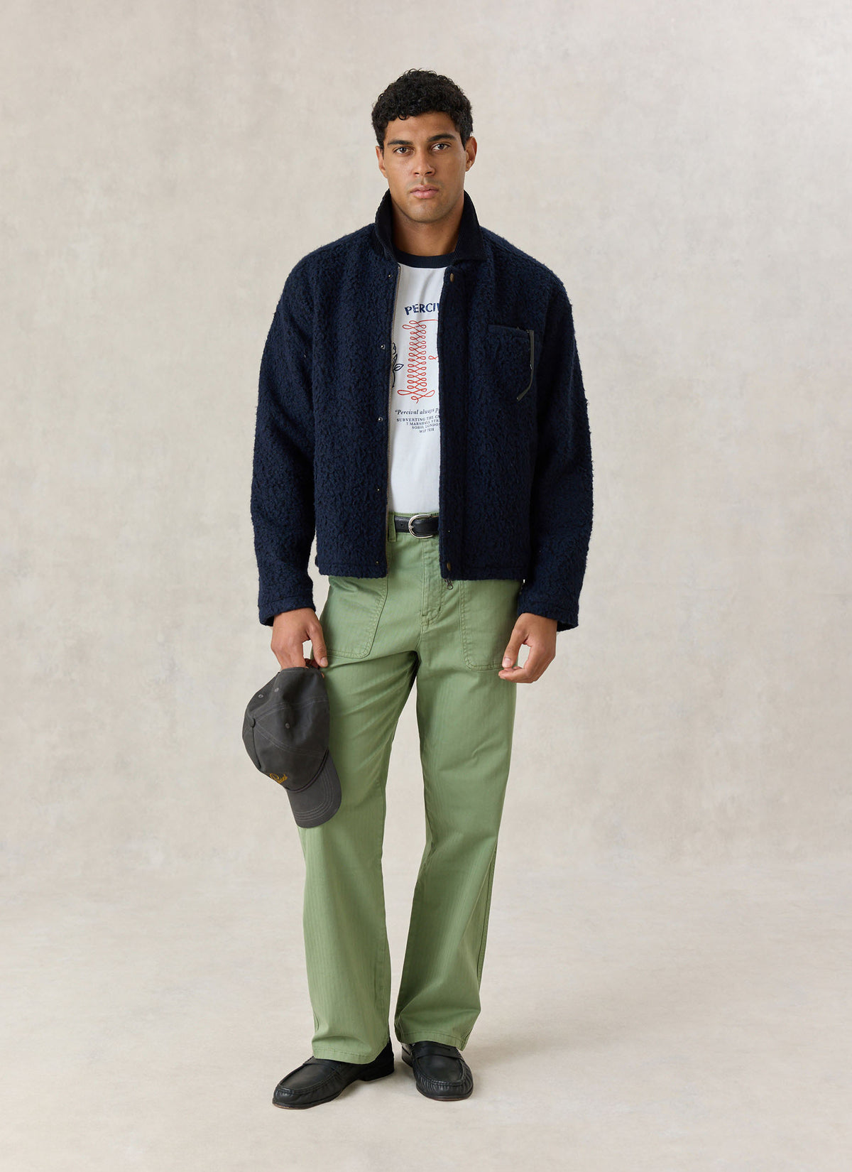Model wearing the navy Percival wool-blend fleece jacket with cord collar, paired with green trousers holding a dark cap.