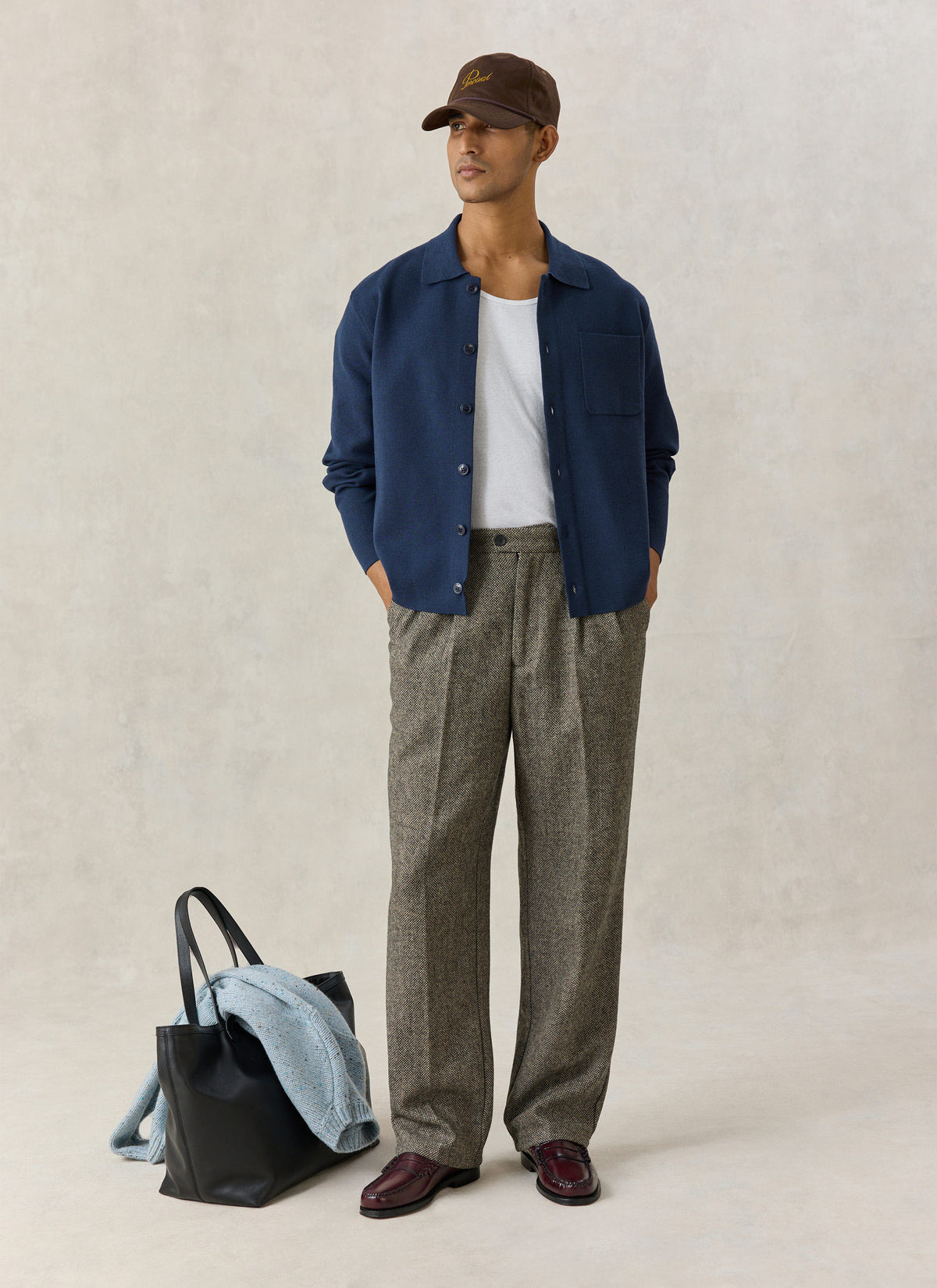 A model wearing the navy Percival Nixon knit collared cardigan open over a white tee, patterned trousers, cap, and loafers.
