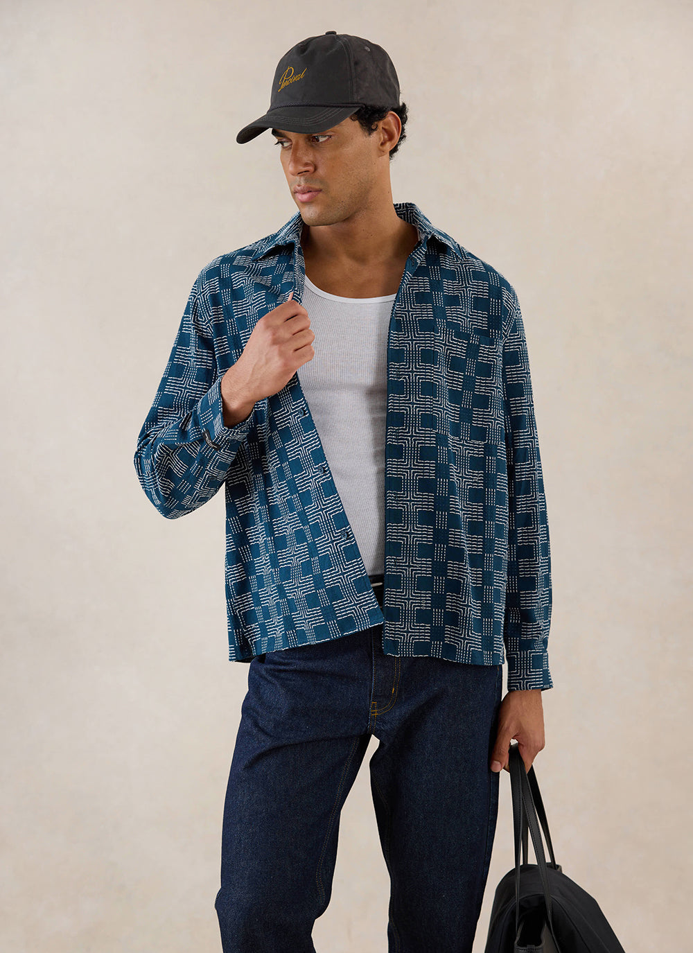 A model wears the navy Percival Nolan Boxy Long Sleeve Shirt over a white vest, styled with dark jeans and a cap.