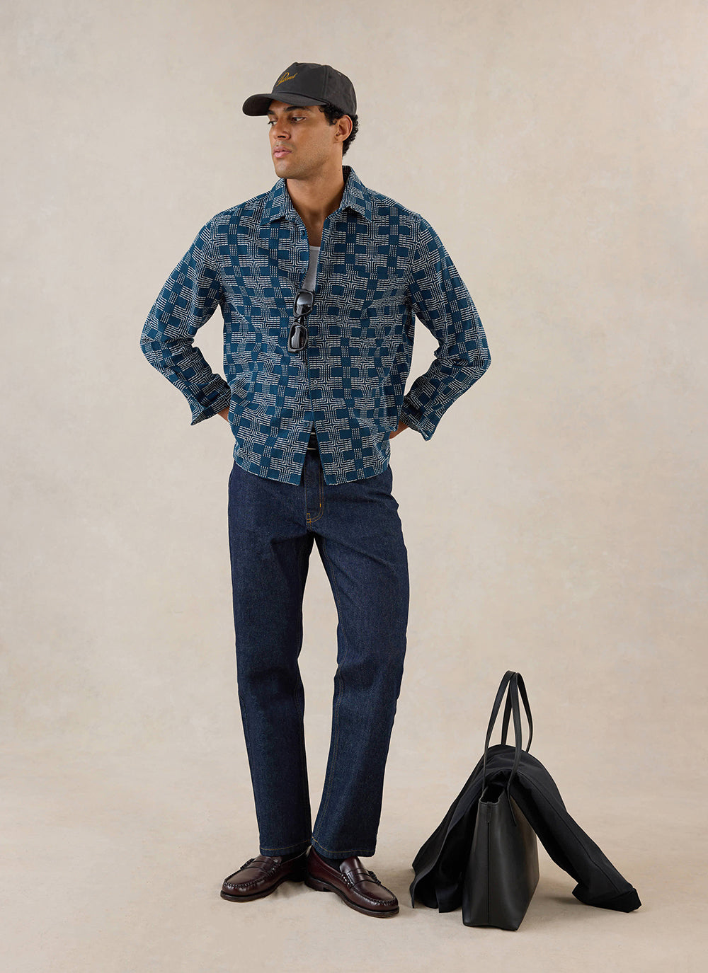A model wears the navy Percival Nolan boxy long sleeve shirt with dark jeans, loafers and a cap in a studio.