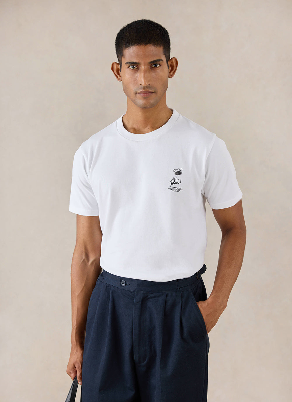 A model wears the white Percival Norman t-shirt, featuring a cocktail chest graphic, styled with dark pleated trousers.