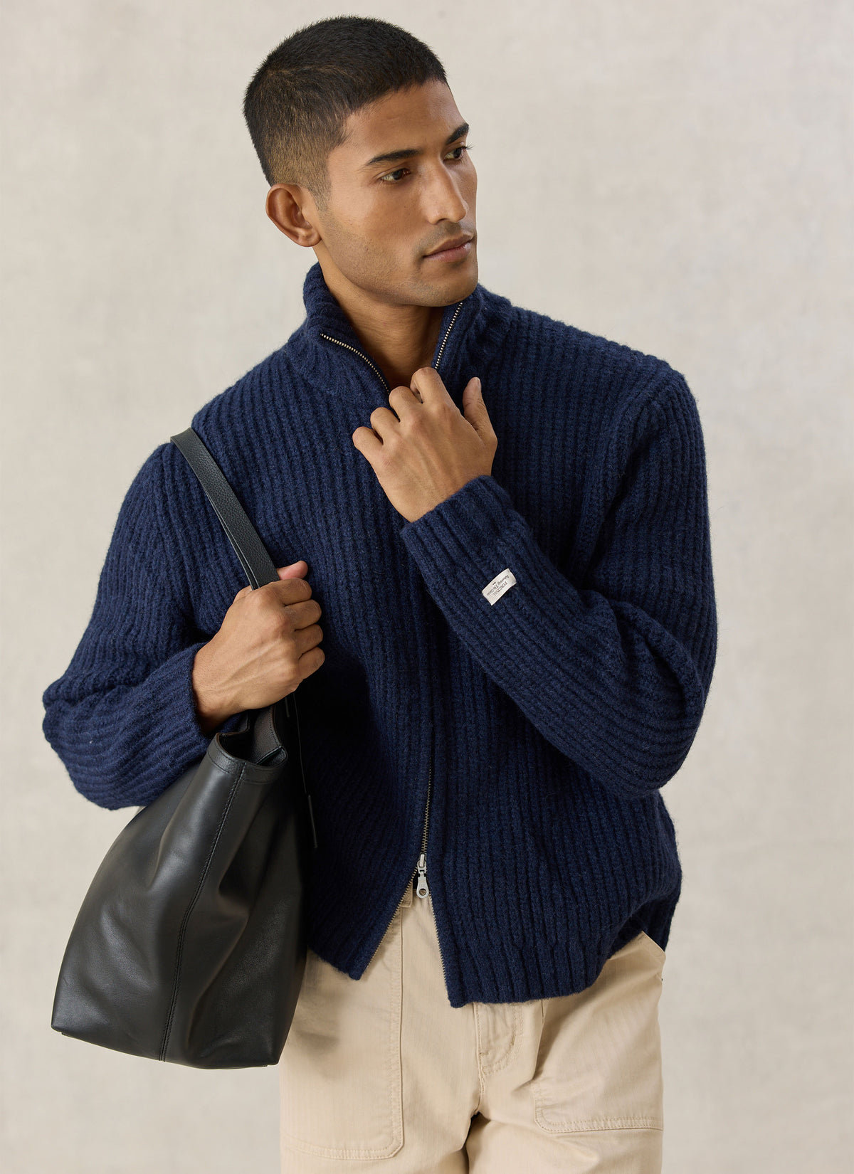 A model wearing the Percival navy Norton ribbed fisherman zip cardigan in alpaca and wool with a black leather tote bag.