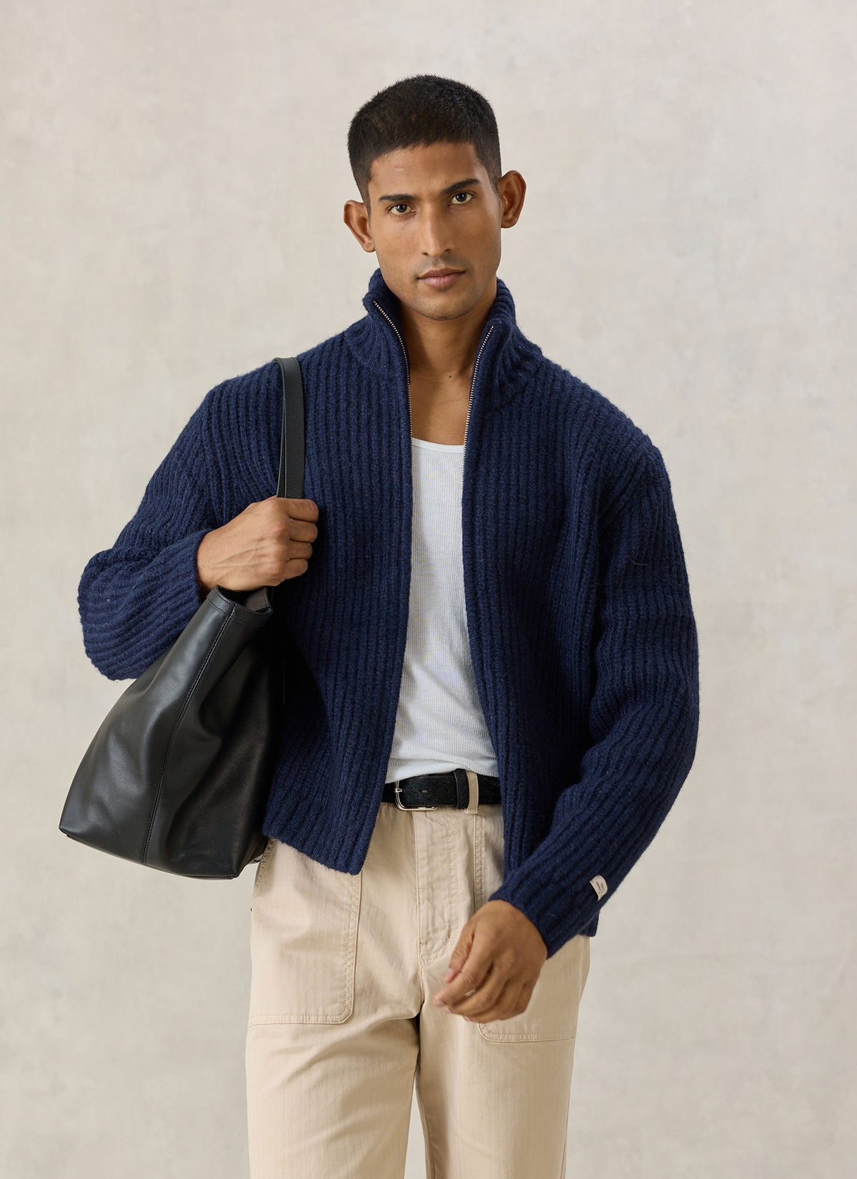 A model wearing the navy Percival Norton ribbed fisherman zip cardigan with a black leather tote bag and beige chinos.