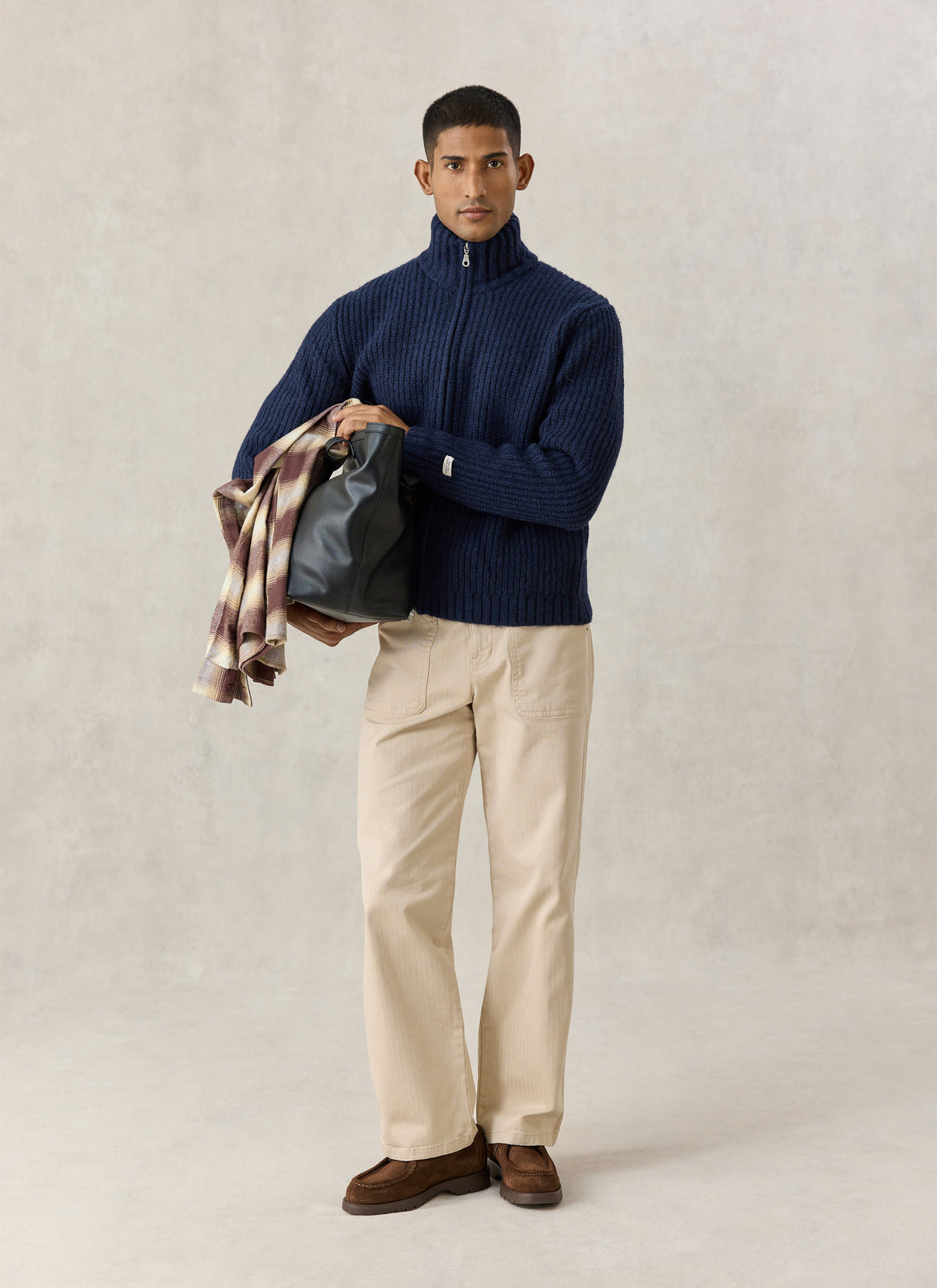 A model wearing the navy Percival Norton ribbed fisherman zip cardigan with beige trousers and holding a black bag.