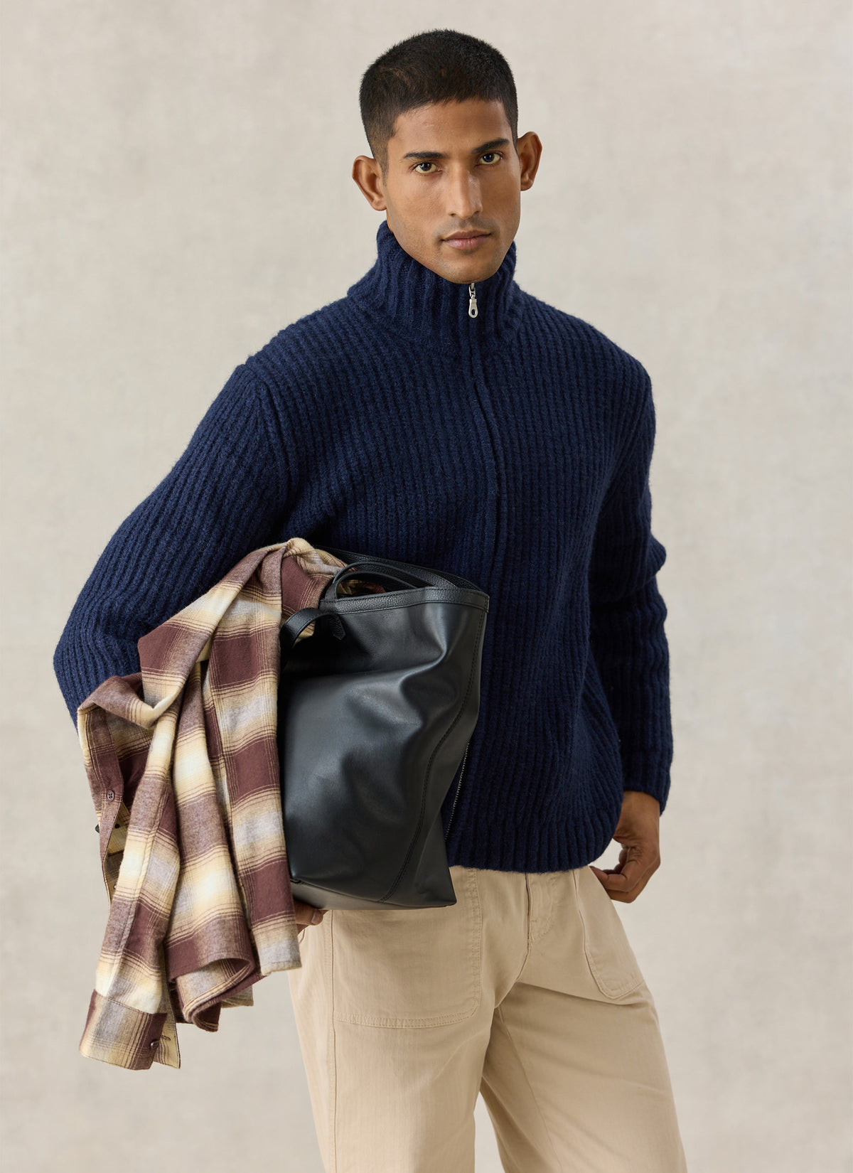 A model wearing the navy Percival ribbed fisherman zip cardigan, holding a black leather bag and plaid shirt.