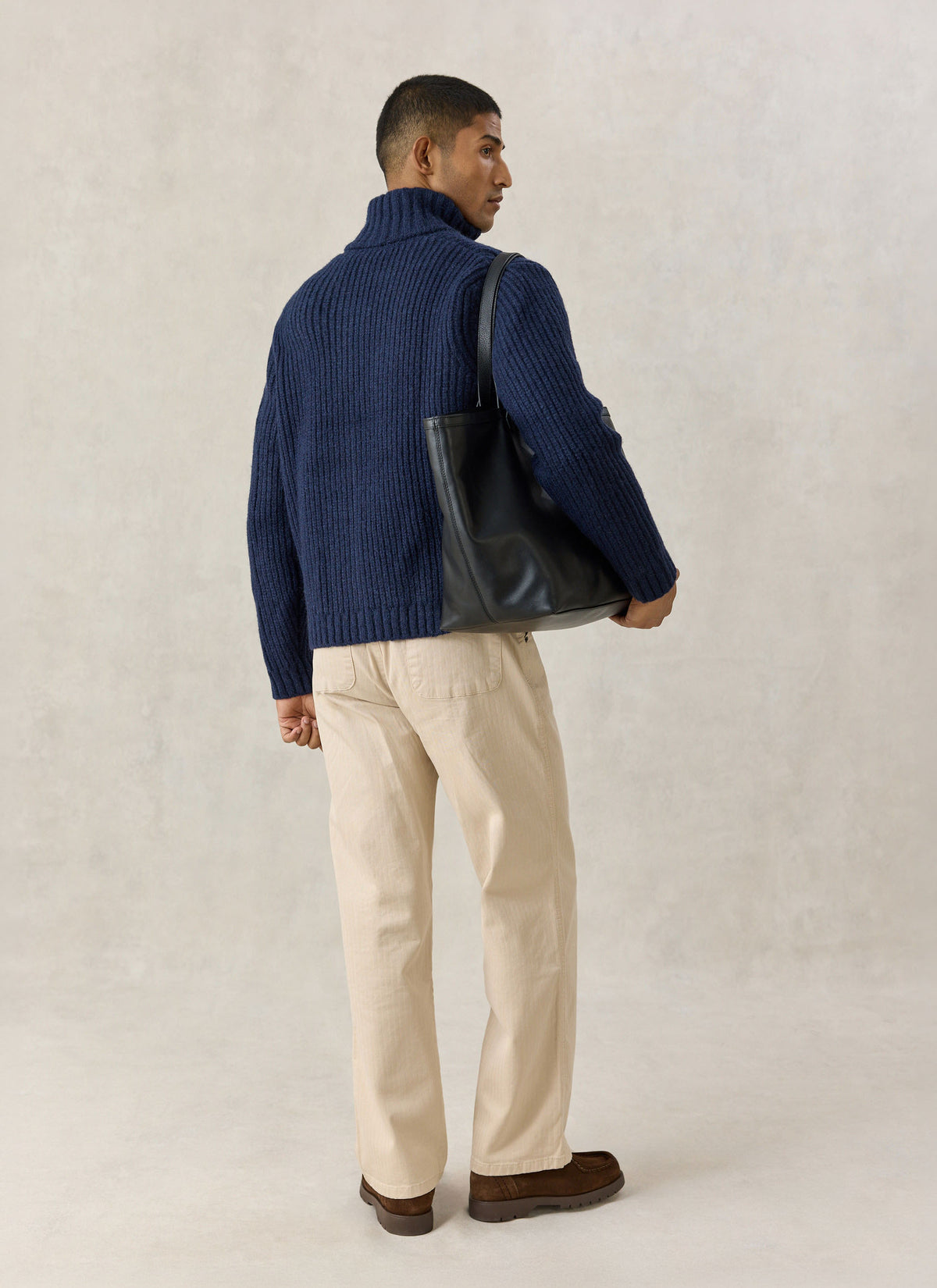 A model wearing the navy Percival ribbed fisherman zip cardigan with a tote bag and beige trousers from behind.