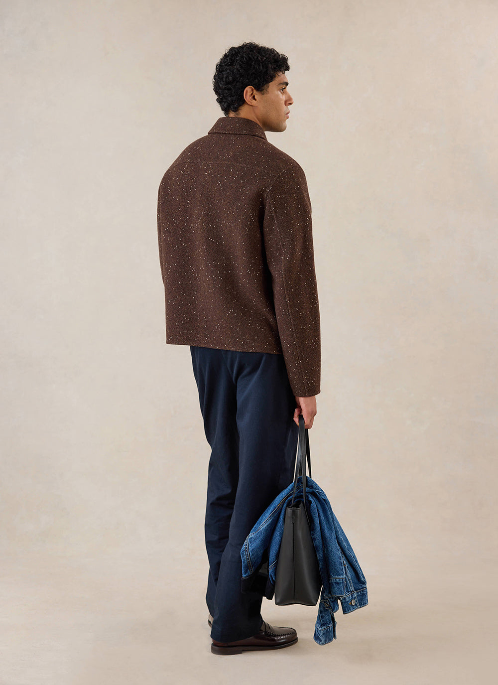 A model stands from behind wearing the brown fleck wool overshirt from Percival, holding a black tote bag in a studio.