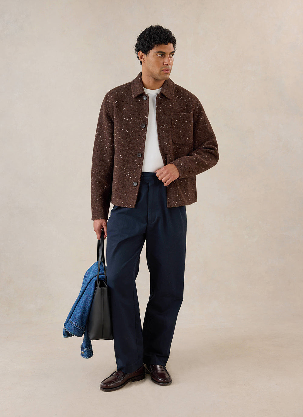 A model wears the brown fleck boxy overshirt from Percival with a white t-shirt, dark trousers, and a black tote bag.