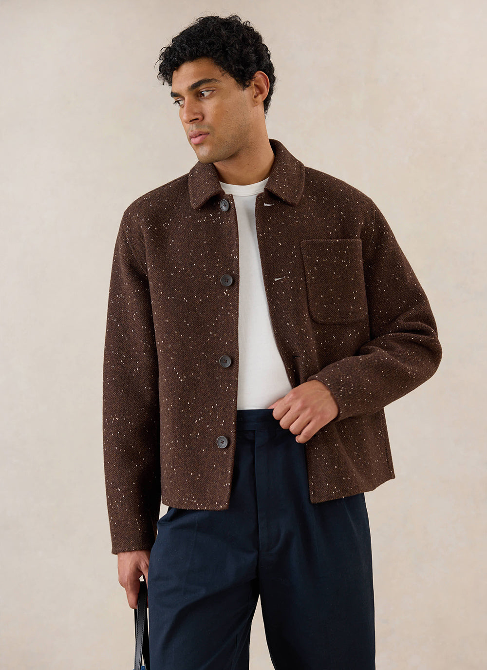 A model wears the brown fleck boxy overshirt from Percival over a white t-shirt and dark trousers in a studio shot.