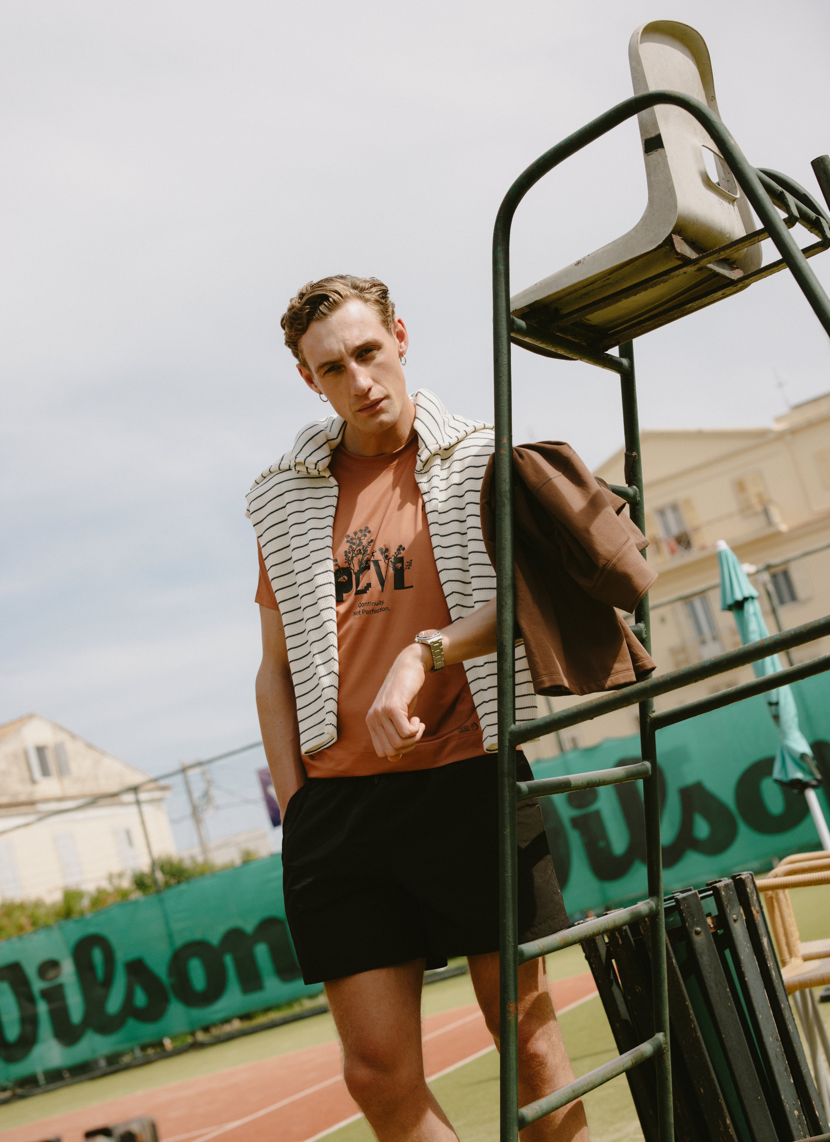 Percival PCVL Graphic Printed Performance Top in rust orange, styled with a striped jumper on a sunny tennis court.