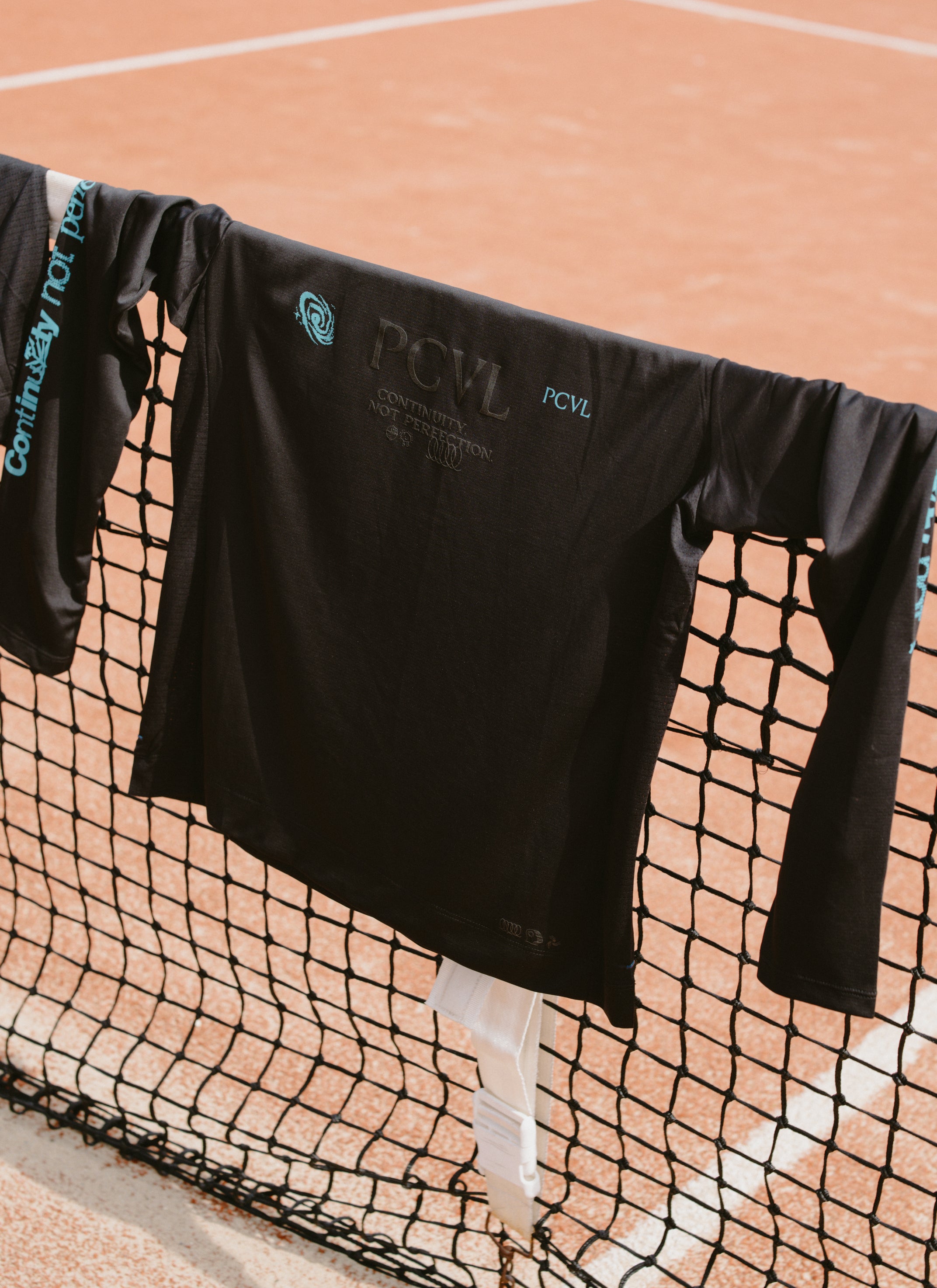 The black Percival PCVL long-sleeve performance top with printed graphics, draped over a tennis net on a clay court.