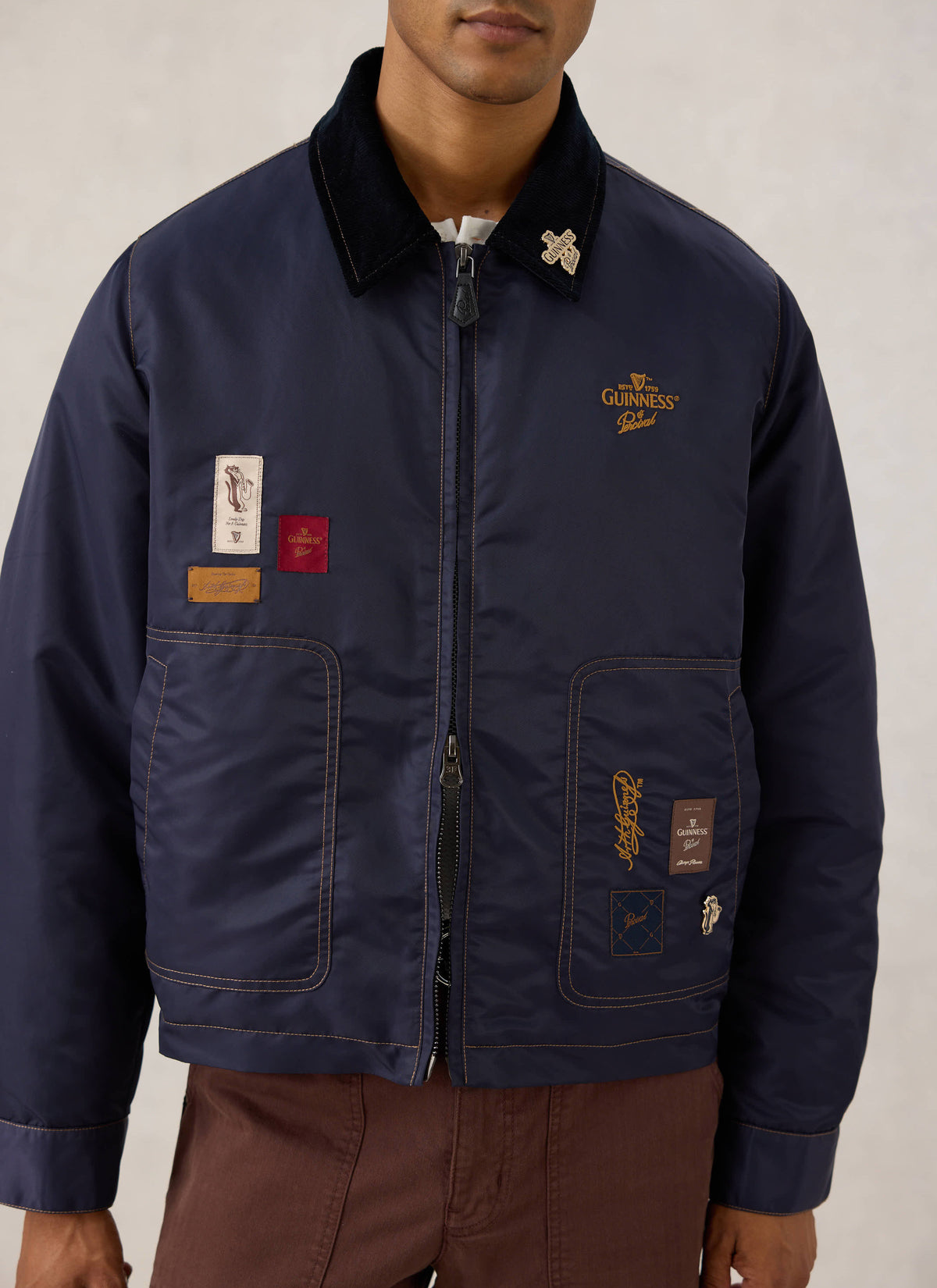 Close-up of the Guinness x Percival navy nylon jacket showing embroidered patches, contrast stitching, and a cord collar.