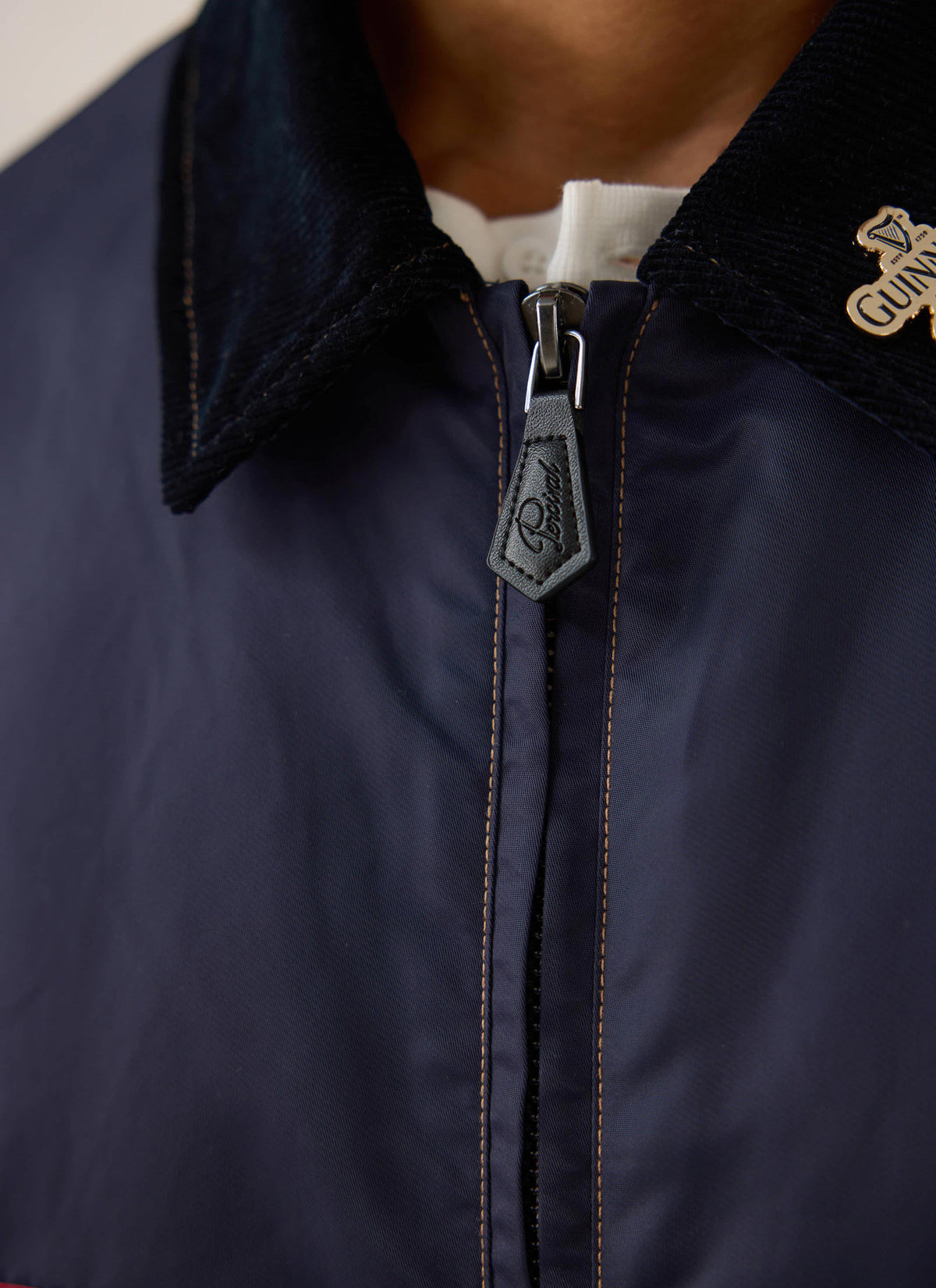 Close-up of the Guinness x Percival navy nylon jacket, showing the cord collar, leather zip pull, and gold pin.