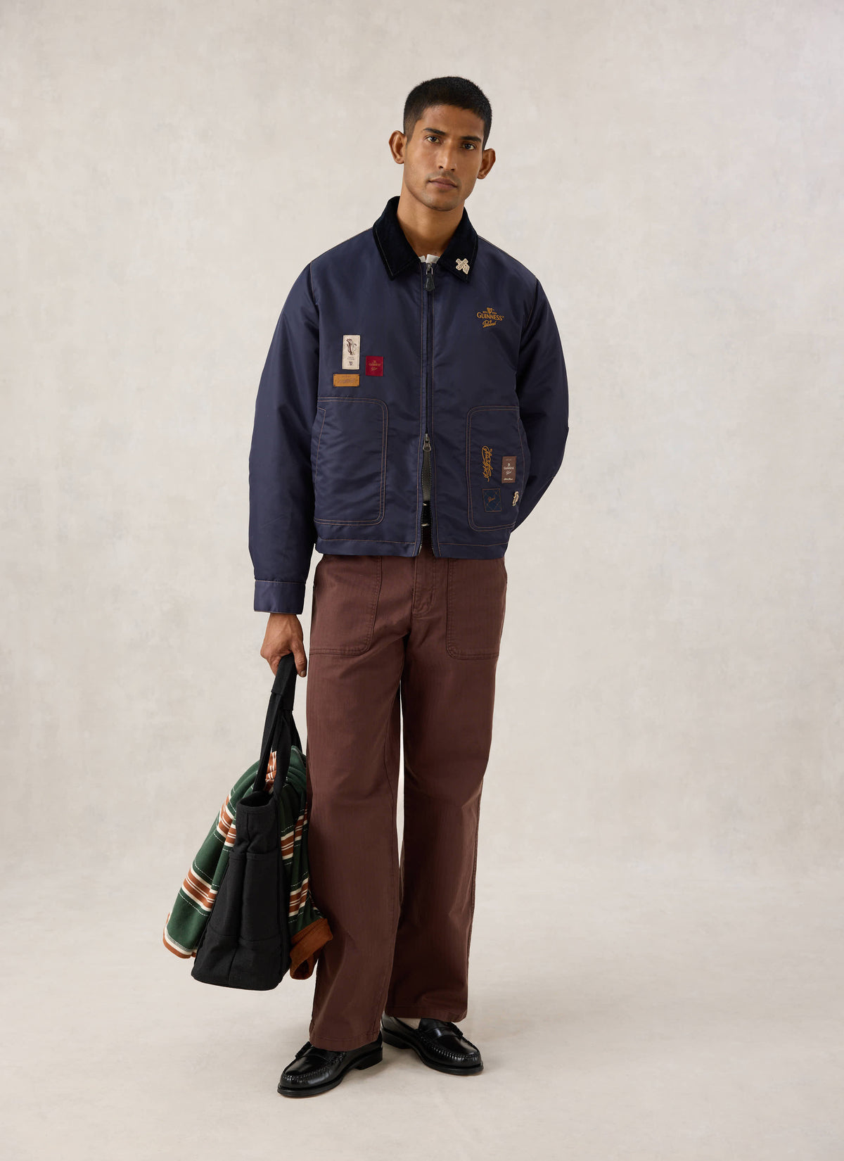 Model wearing the Guinness x Percival navy nylon jacket with embroidery and patch details, brown trousers, and a tote bag.