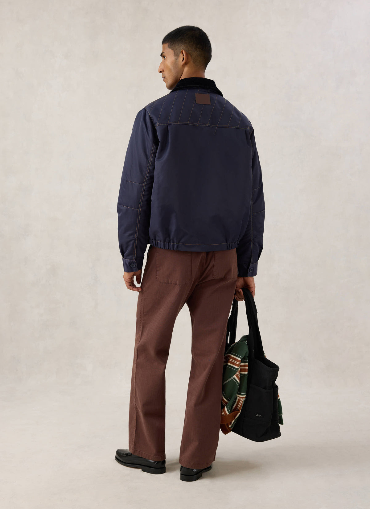 Rear view of a model wearing the Guinness x Percival navy nylon jacket, styled with brown trousers and a bag.