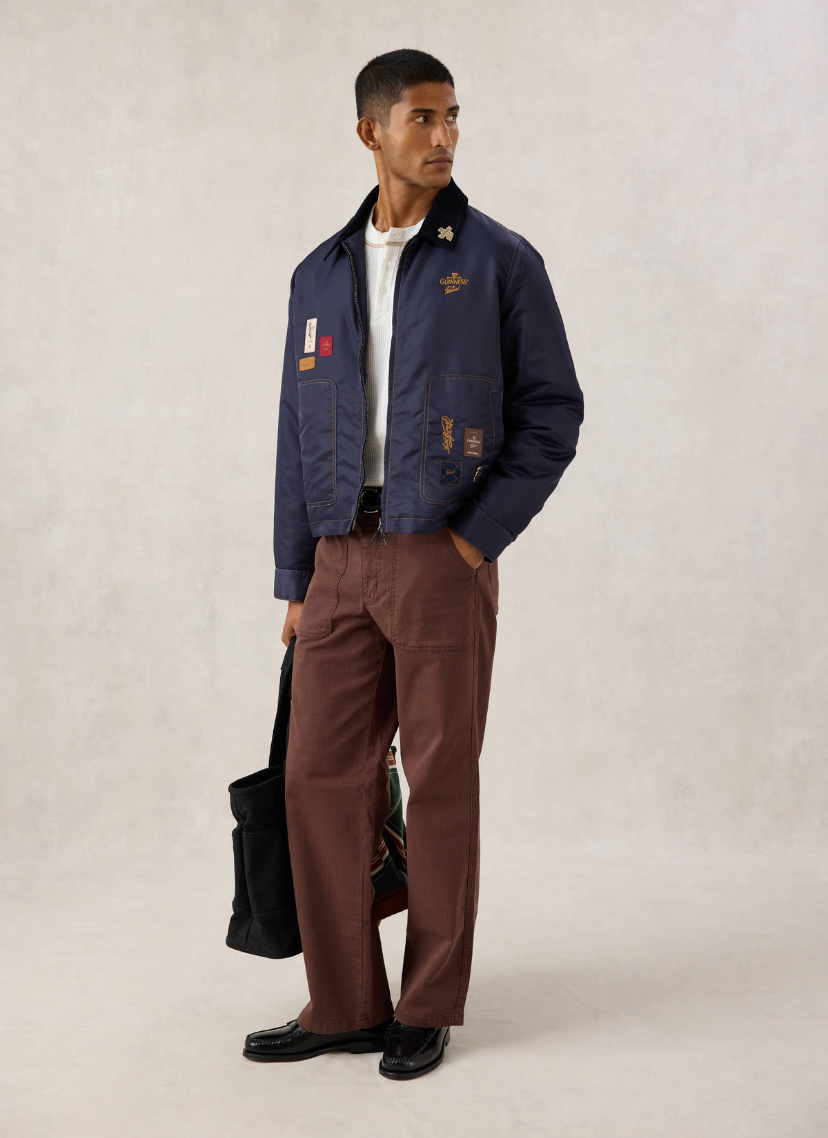 Model wearing the Guinness x Percival navy nylon jacket with embroidery and patch details, paired with brown trousers.