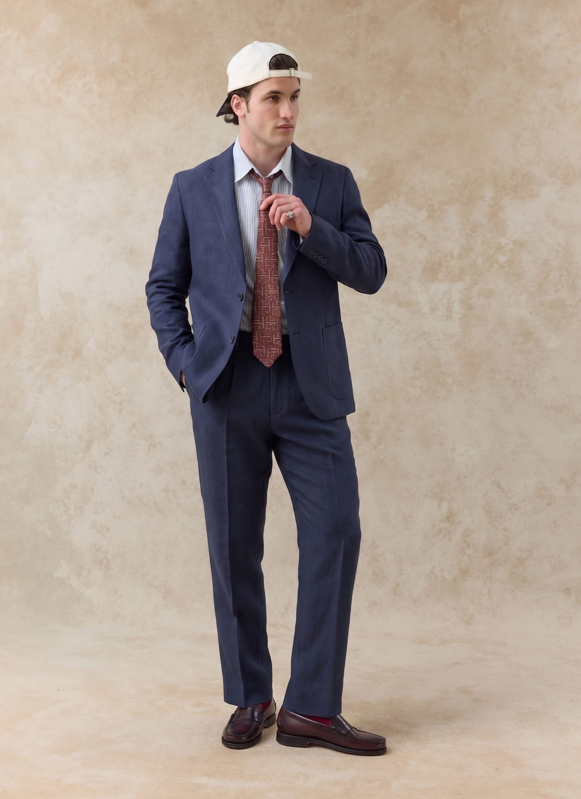 Navy blue Percival tailored linen blazer and matching trousers, styled with a striped shirt and white baseball cap.