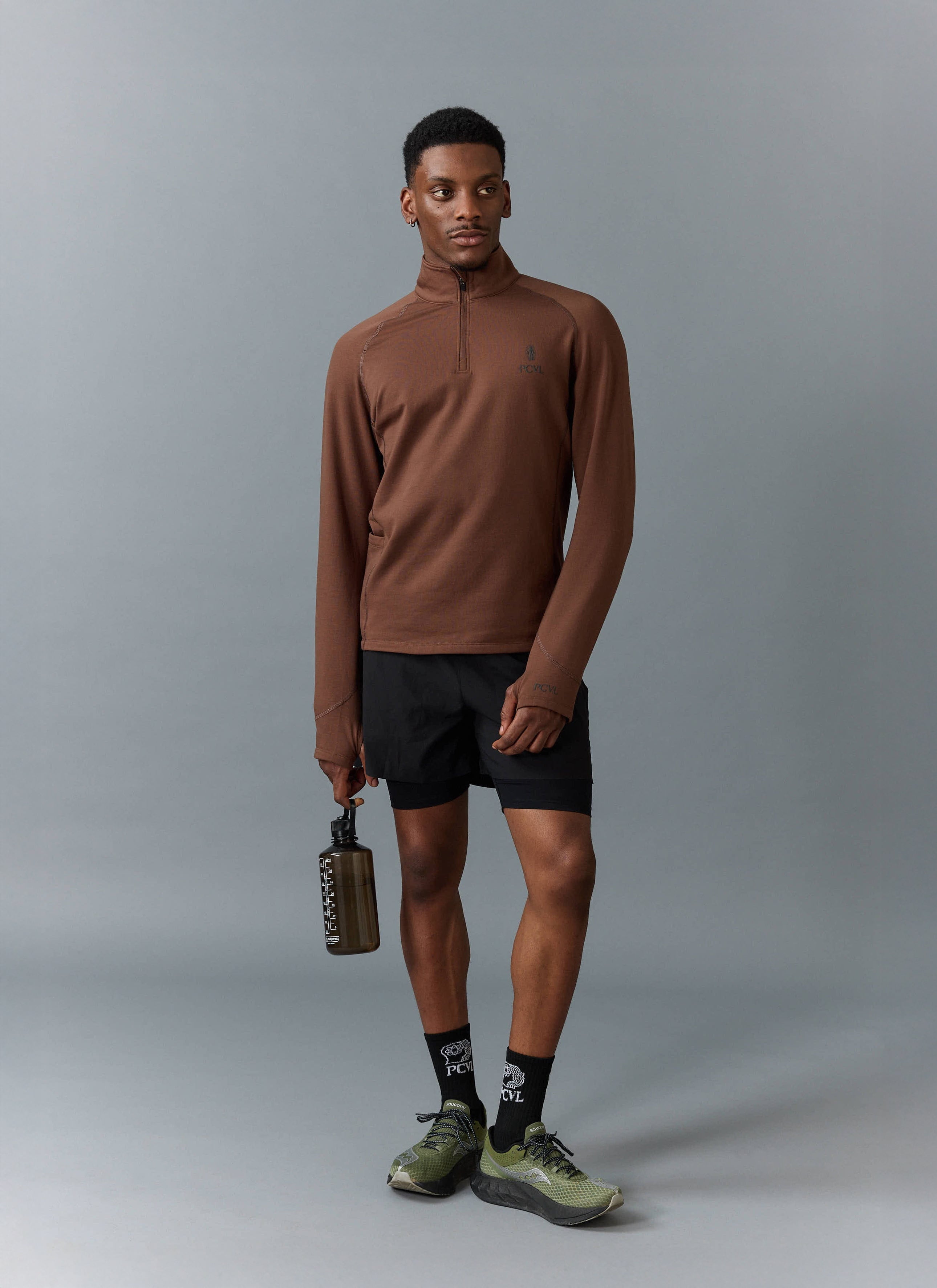 A model wears the Percival PCVL Base Layer Quarter Zip in rust, styled with black activewear shorts and trainers.
