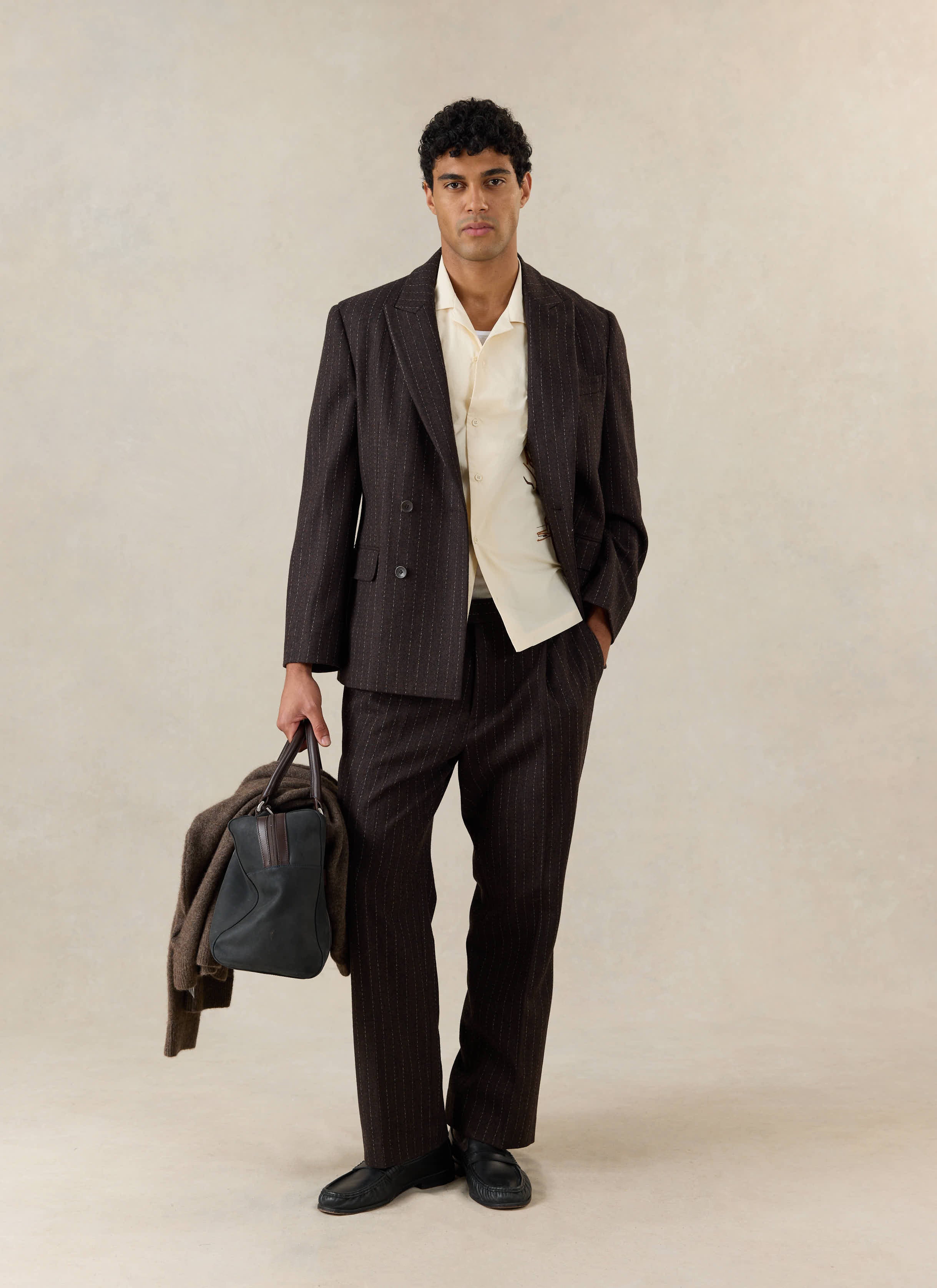 A model wears the cream Percival Cuban shirt under a brown pinstripe suit, holding a dark bag and jacket, with black loafers.