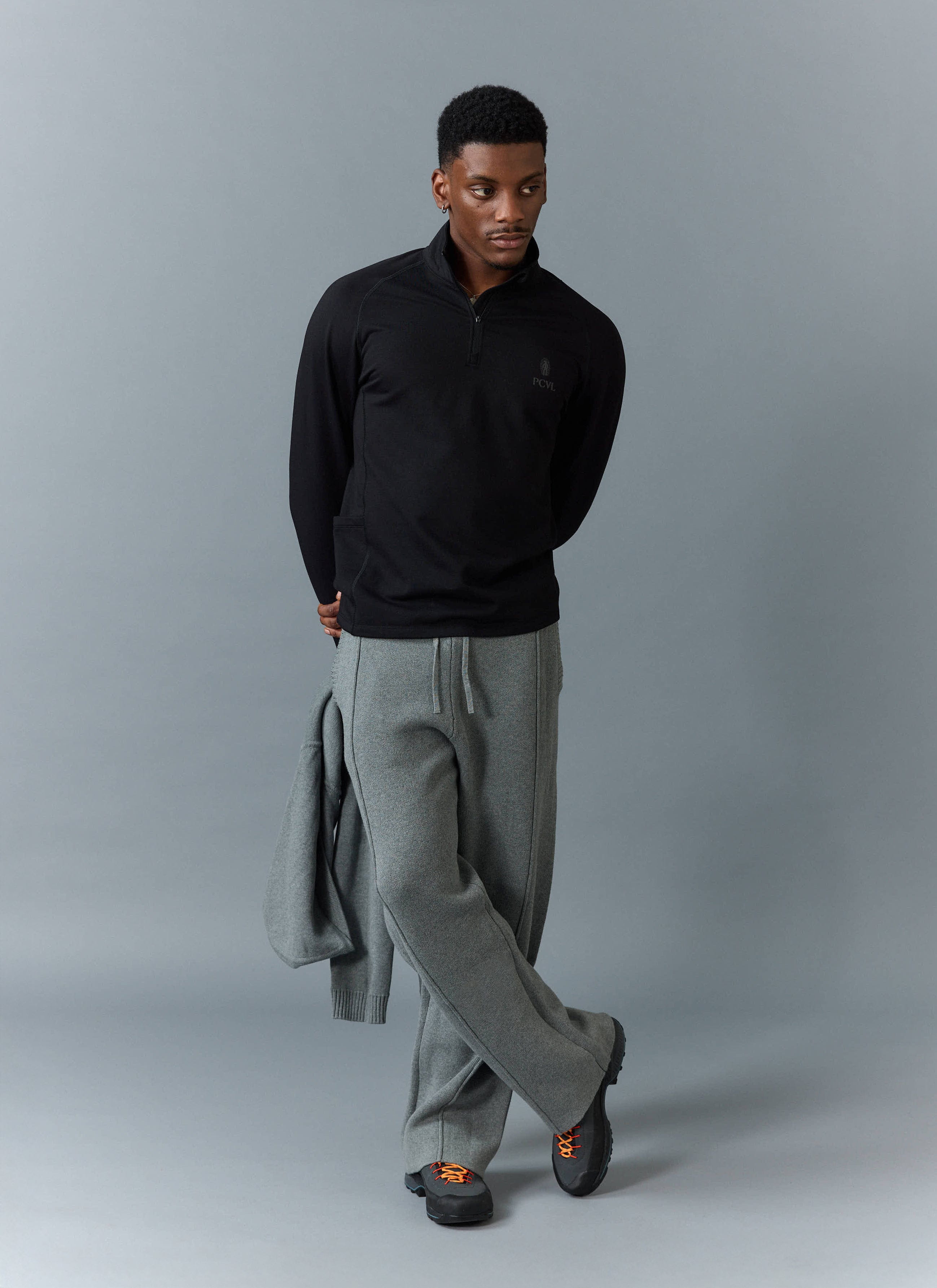 A model wears the black PCVL Base Layer Quarter Zip Jacket from Percival, styled with grey trousers and black walking shoes.